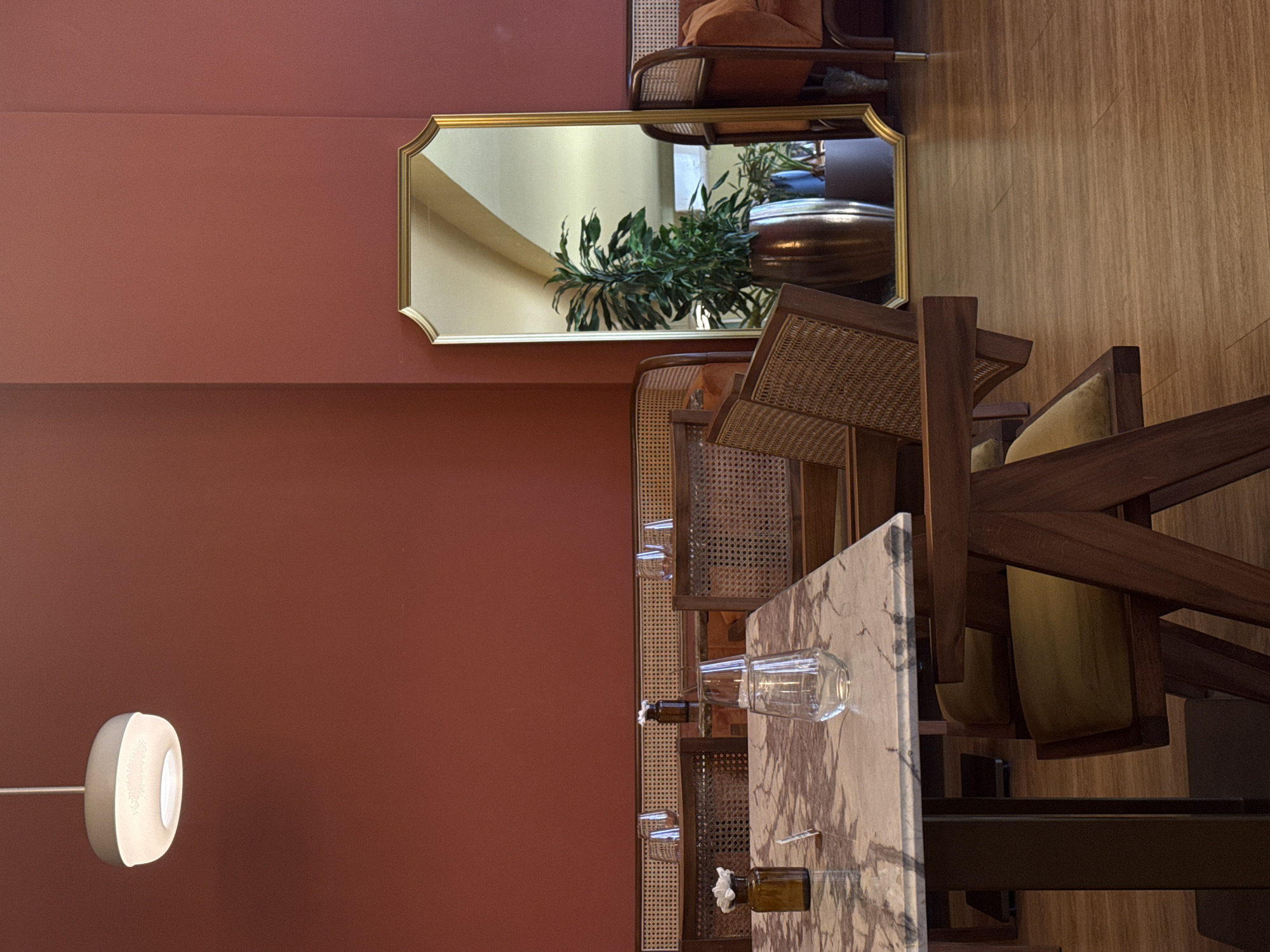 The image depicts a room setting, likely in a restaurant or hotel, in Boreti, Montenegro. The interior features a terracotta-colored wall on the left, complemented by wooden furniture, including a table with a marble top and chairs with woven rattan backs. A gold-framed mirror on the wall reflects an interior view of the room, including a plant and decorative vase. A modern-style lamp is visible on the left wall. Glassware and small decorative bottles are placed on the table, suggesting a setup for dining. The floor is made of light-colored wood. The scene is still, with no people visible.