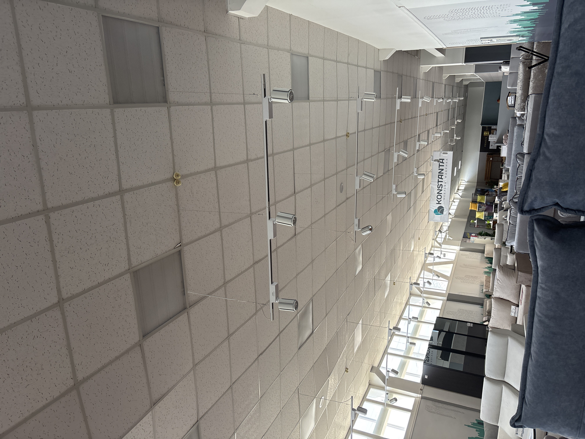 The image shows the interior of a furniture store, possibly a showroom, with a view of the ceiling and aisles of furniture. The ceiling is made of acoustic tiles with a grid system, and suspended track lighting with multiple spotlights is visible.
The scene is oriented such that the camera is looking upwards towards the ceiling and then downwards into the store space. Along the ceiling, there are several modern, cylindrical spotlights mounted on thin metal rods, connected by wires that span across the space.
In the background, furniture items such as sofas, armchairs, and cushions are displayed in rows, creating aisles. Some of these are large, upholstered pieces in various neutral tones like gray and beige. There are also some decorative elements on the walls, including a graphic design that appears to be stylized trees or a cityscape. A prominent banner hangs from the ceiling with the text "KONSTANTA" and "МЕБЛЕВА ФАБРИКА" (Furniture Factory) in Cyrillic script.
The lighting suggests it could be daytime, as natural light seems to be entering from large windows visible in the background, casting a bright glow on the furniture. The overall impression is of a well-lit, modern retail environment. The image is taken from a low angle, looking upwards, which emphasizes the scale of the space and the overhead lighting system. There are no people visible in the frame. The location is identified as Zaporizhzhia, Ukraine.