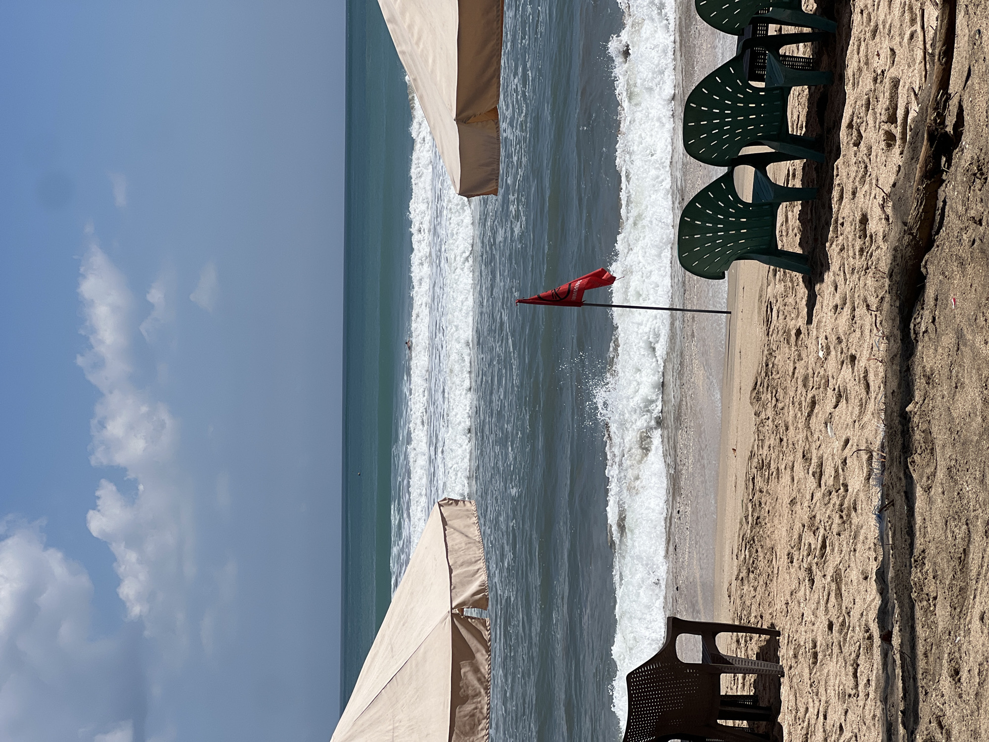 The image captures a beach scene in Kuta, Indonesia, under a bright, partially cloudy sky. The foreground features a sandy beach with several plastic chairs, some dark brown and others dark green. A fallen log lies in the sand in the lower right portion of the frame. In the middle ground, a red flag is planted in the sand, signaling caution or a warning. Two large, light-colored beach umbrellas are partially visible on the left and right sides, providing shade. The ocean in the background displays moderate waves with white foam breaking near the shore. A person is visible in the distance, swimming or wading in the water. The environment is characteristic of a tropical beach resort.