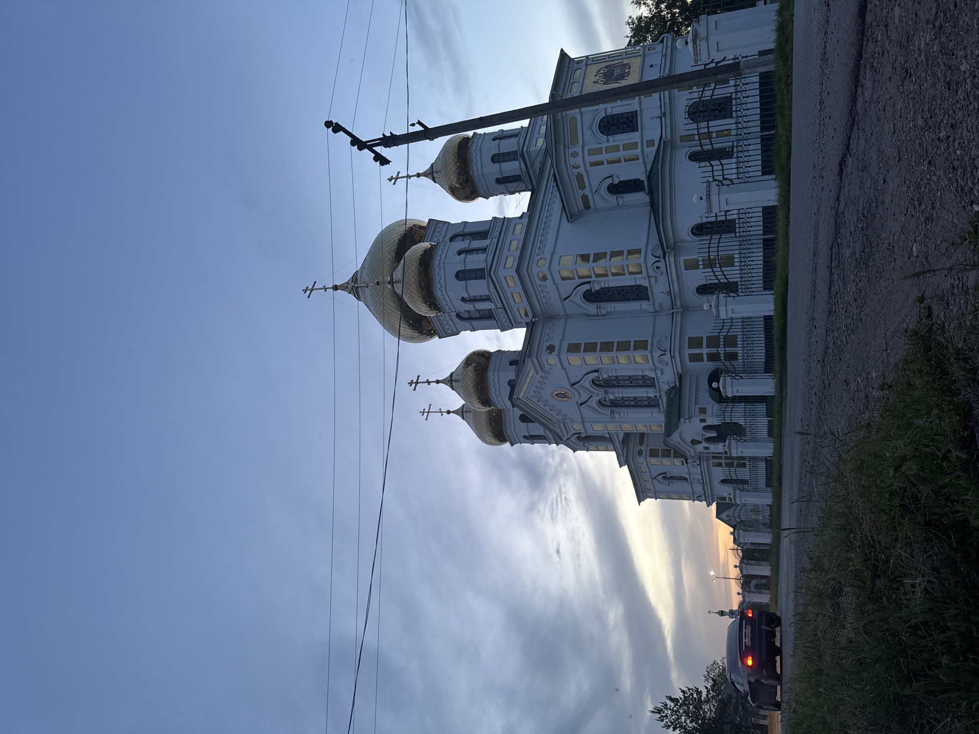 The image depicts the Cathedral of the Nativity of the Blessed Virgin in Novodonetskaya, Russia. The cathedral is a white building with golden domes. The photograph is taken from the side of the road. A black car is parked on the side of the road further down. Electrical wires extend across the upper left portion of the image. The sky is a mix of blue and grey, with a streak of sunset orange visible in the distance.