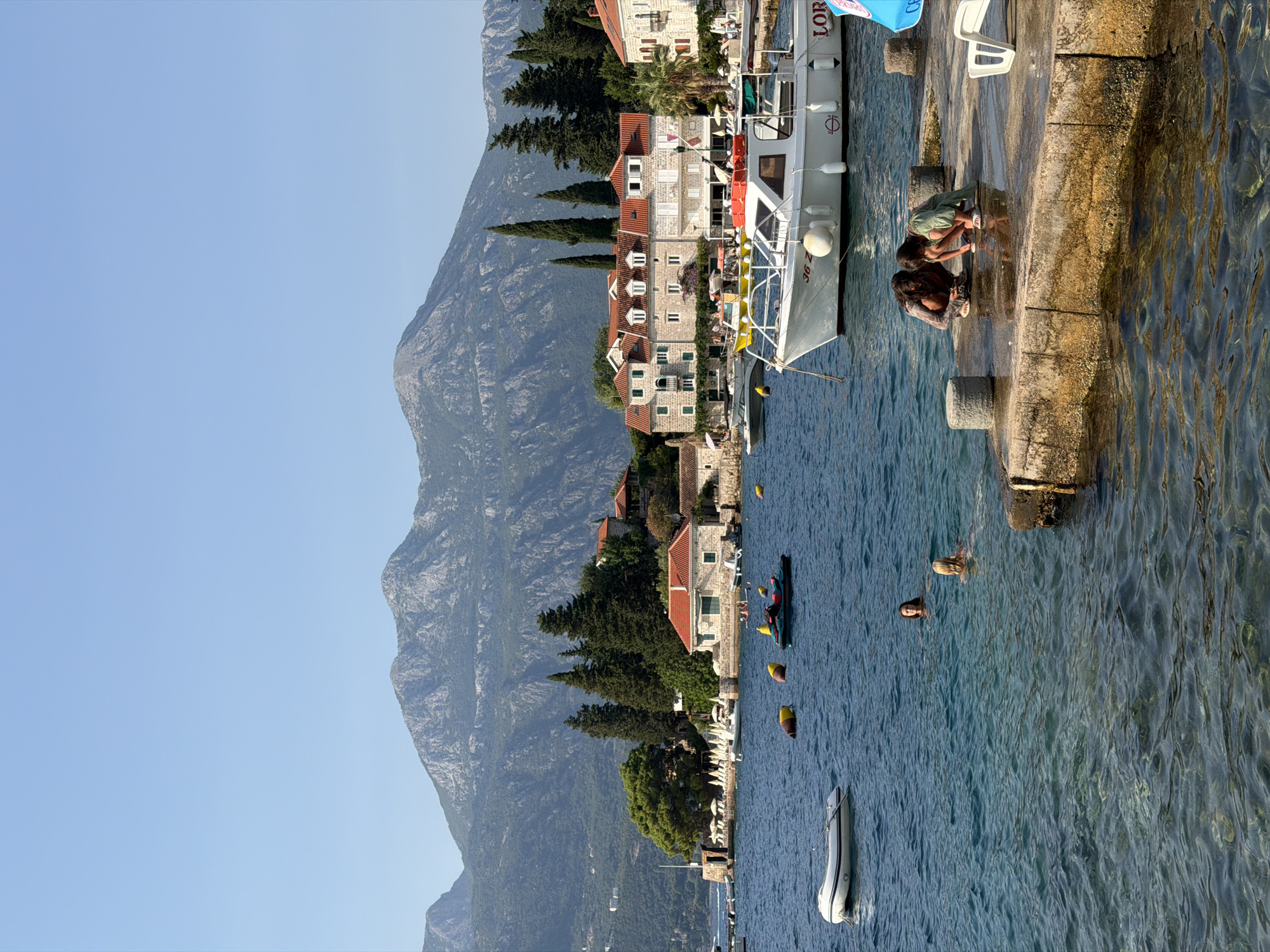 This is a vertical, eye-level, outdoor, daytime, full shot of a coastal scene in Zelenika, Montenegro. In the foreground, the clear, blue water laps against a stone pier where two children, one with dark hair and the other with light hair, are crouched down, interacting with something on the pier. To the left of the pier, two women are swimming in the water. A white inflatable boat is moored to the left of the pier. In the middle ground, several boats are anchored in the water. A white boat with a red and white stripe is prominently featured. Behind the boats and pier, a town with white and red-roofed buildings lines the shore. Lush green trees and palm trees dot the landscape. In the background, a large, rugged mountain range with rocky peaks rises under a clear blue sky. The overall atmosphere is peaceful and sunny, suggesting a vacation or leisure setting.