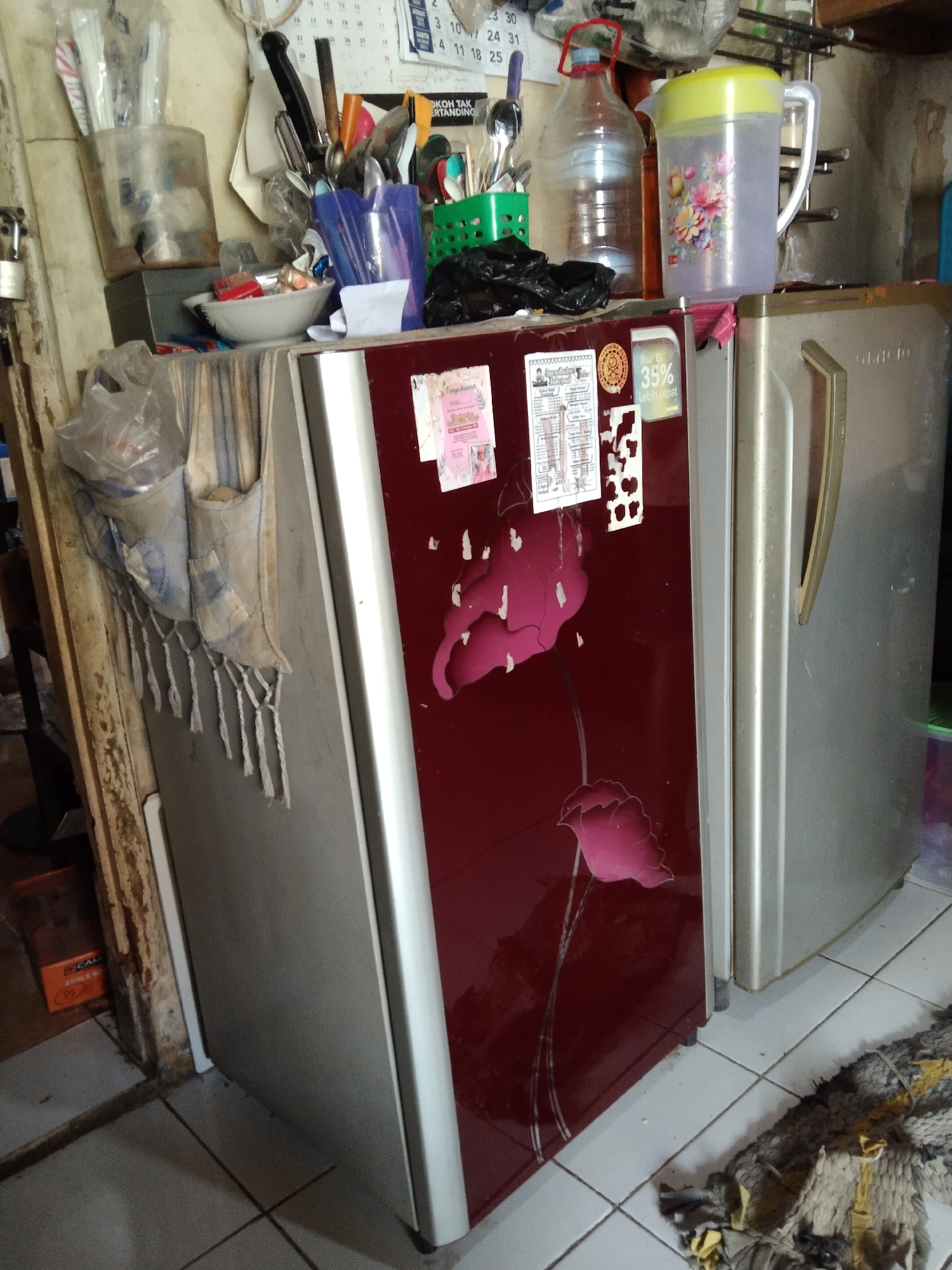 The image depicts a cramped interior space, likely a kitchen or food preparation area, in Parung, Indonesia. The focal point is a maroon-colored refrigerator with a floral design, situated next to a silver-colored refrigerator. Various kitchen utensils, including spoons and knives, are visible in containers on top of the maroon refrigerator. A large plastic water jug and a smaller jug with a floral pattern are also present. Other objects include a white bowl with what appears to be food items, a black plastic bag, and various papers attached to the refrigerator doors. The floor is tiled, and a disheveled rug or cloth lies on the floor in the foreground. The overall impression is one of a functional but cluttered domestic environment.