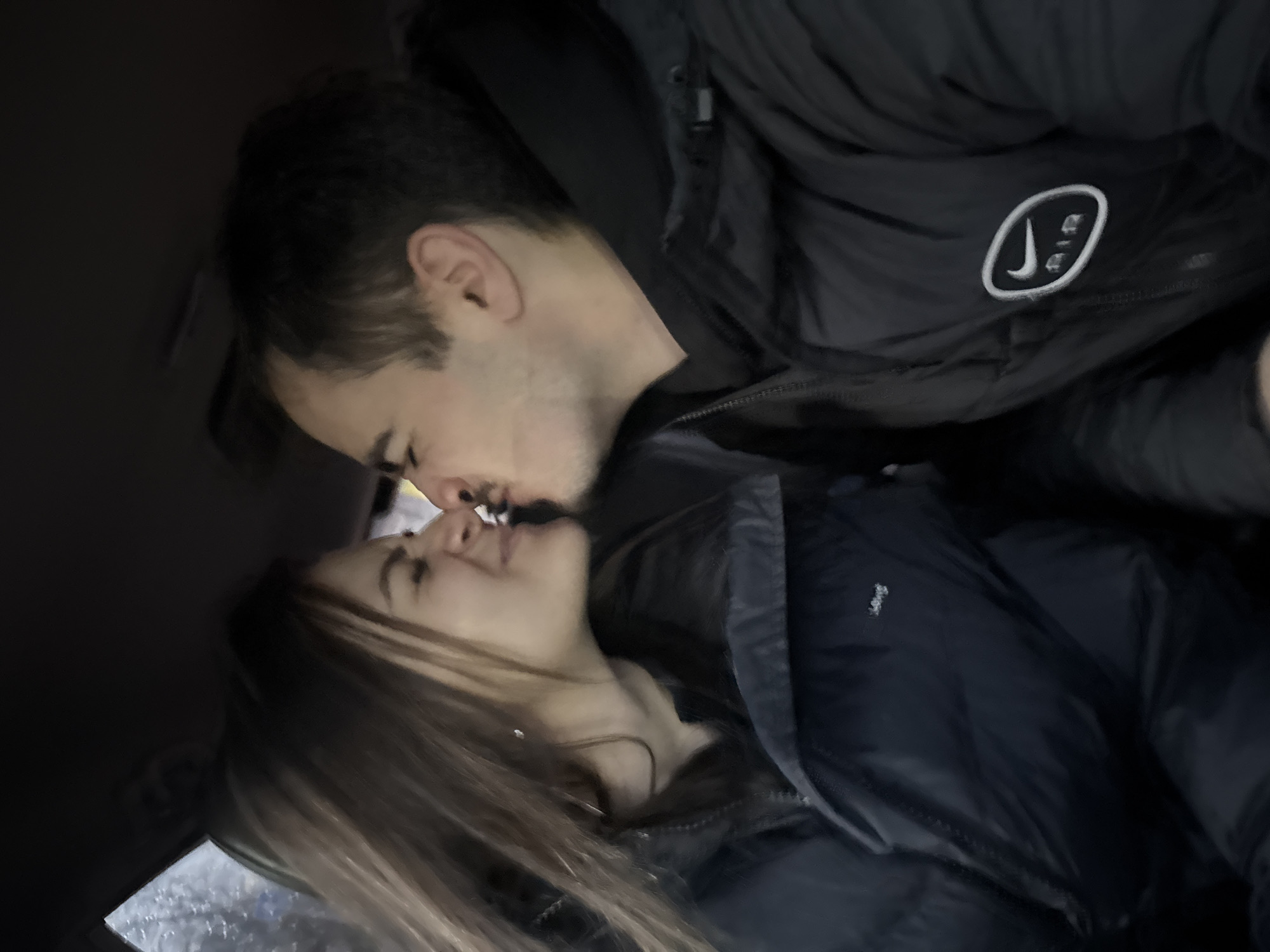 A man and a woman are kissing. Both are wearing dark jackets. The man's jacket has a white circular logo with what appears to be an altered Nike swoosh and "AIR" written inside the circle. The photo appears to have been taken inside a car. The location cannot be determined from the image.