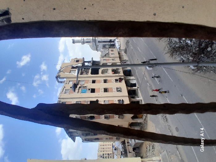 The image depicts a damaged urban street scene in Khan Yunis, Palestinian Territory, viewed through what appear to be two thick, dark, corroded rebar or concrete fragments in the immediate foreground, with a pockmarked, light-colored wall section visible on the far right. In the mid-ground, an asphalt road with white lane markings spans horizontally. Approximately eight children, appearing to be young males, are scattered across the street, engaged in playing soccer with a light-colored ball. One child wears a red shirt, another a yellow shirt. A grey streetlamp pole stands to the right of the children. Along the right side of the road, piles of rubble and debris are visible.
In the background, a multi-story residential building with light-colored walls and dark brown decorative elements is severely damaged, exhibiting large sections of missing walls and roof, exposed rebar, and broken windows. Laundry hangs from several balconies. To the right of this building, a mosque with a tall, slender minaret and a large dome is visible, also showing signs of structural damage. Further left in the background, other multi-story residential buildings, some appearing less damaged, line the street. The sky above is clear blue with scattered white clouds.