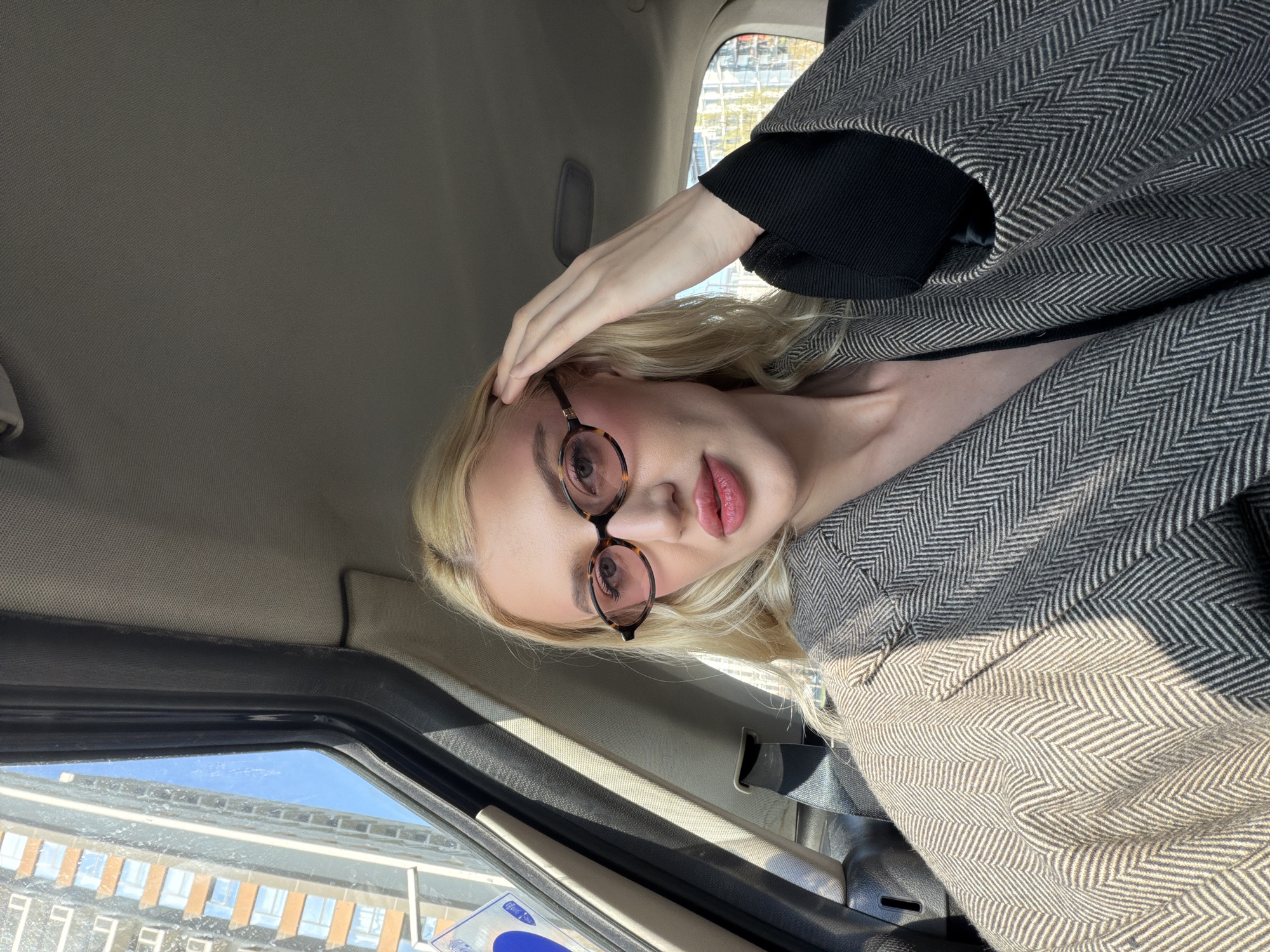 A blonde-haired woman with glasses poses for a photo in a car, wearing a herringbone coat. The background shows buildings outside the car window, typical of an urban environment, with a glimpse of a blue and white parking sign. This is likely a street scene. Given the prompt information, the location is identified as Belgrade, Serbia.