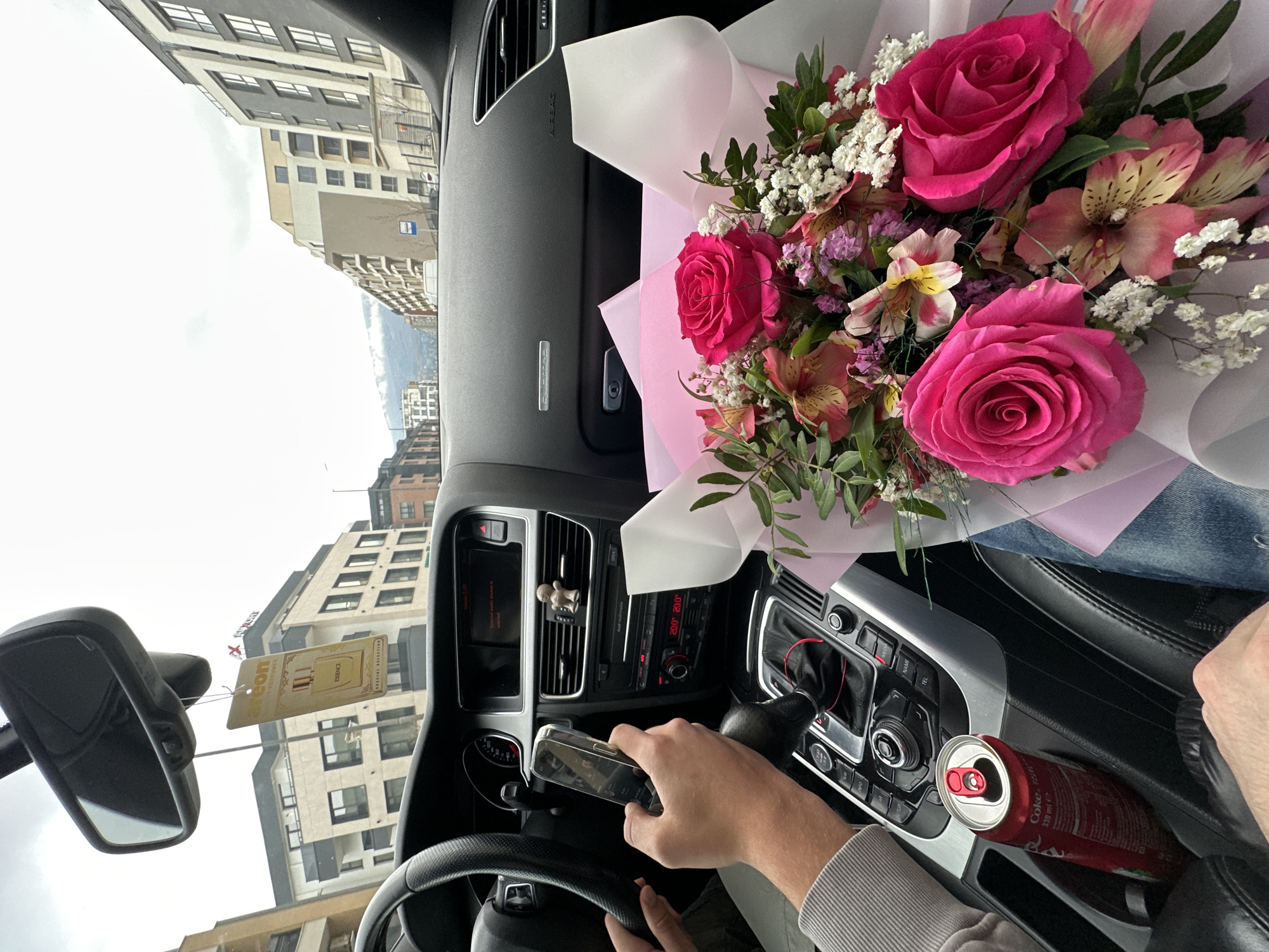 A first-person, upside-down view from inside an Audi car shows a bouquet of pink roses and other flowers on the passenger seat. The driver is holding a mobile phone with a reflective surface. A can of Coca-Cola sits in the cupholder. The car's dashboard, steering wheel, and rearview mirror are visible. Outside, buildings and a cloudy sky are seen. The location is Sofia, Bulgaria.