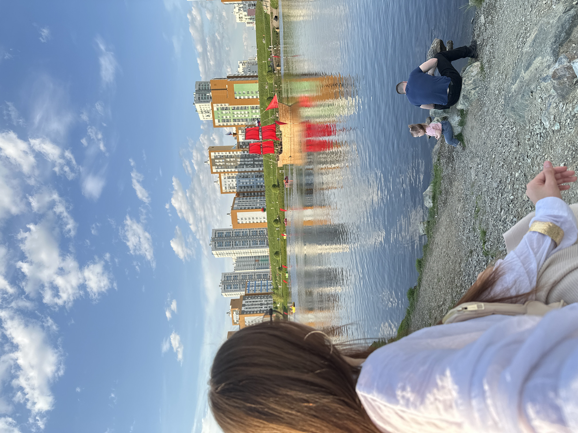 A woman with long brown hair, wearing a white shirt and beige jacket, stands near a body of water, possibly a lake or river, in Yekaterinburg, Russia. On the opposite bank of the water are several tall buildings with a variety of colors, including beige, white, and orange. The buildings reflect in the water. A structure resembling a ship with red sails floats in the water near the shore. To the right of the woman, a man and a young girl sit on a rocky shore near the water's edge. The man wears a dark blue shirt and dark pants. The girl is wearing a pink shirt and jeans. The sky is blue with scattered white clouds. The photo is taken from the woman's perspective, with her back and hand visible in the foreground.