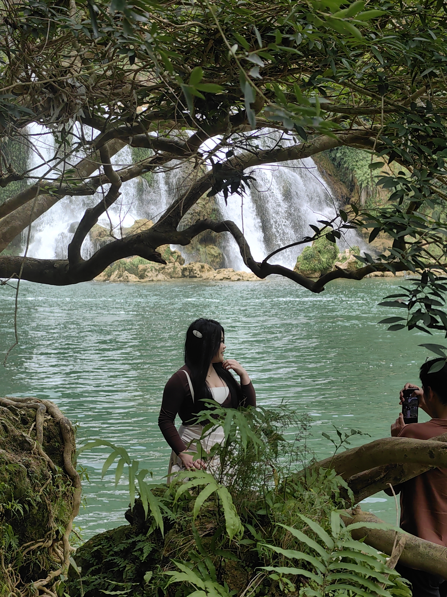 A medium shot photograph taken in Daxin County, China depicts a woman standing in a green body of water. She is wearing a dark brown, long-sleeved jacket over a white top and a light-colored skirt. Her dark hair is pulled back. She is looking to her right, her right hand raised to her face. Behind her is a waterfall cascading over rocks. A tree branch frames the top of the waterfall. The foreground features foliage. To the right of the woman, a person is partially visible, holding a mobile phone up, as if taking a picture. The water is calm with ripples.