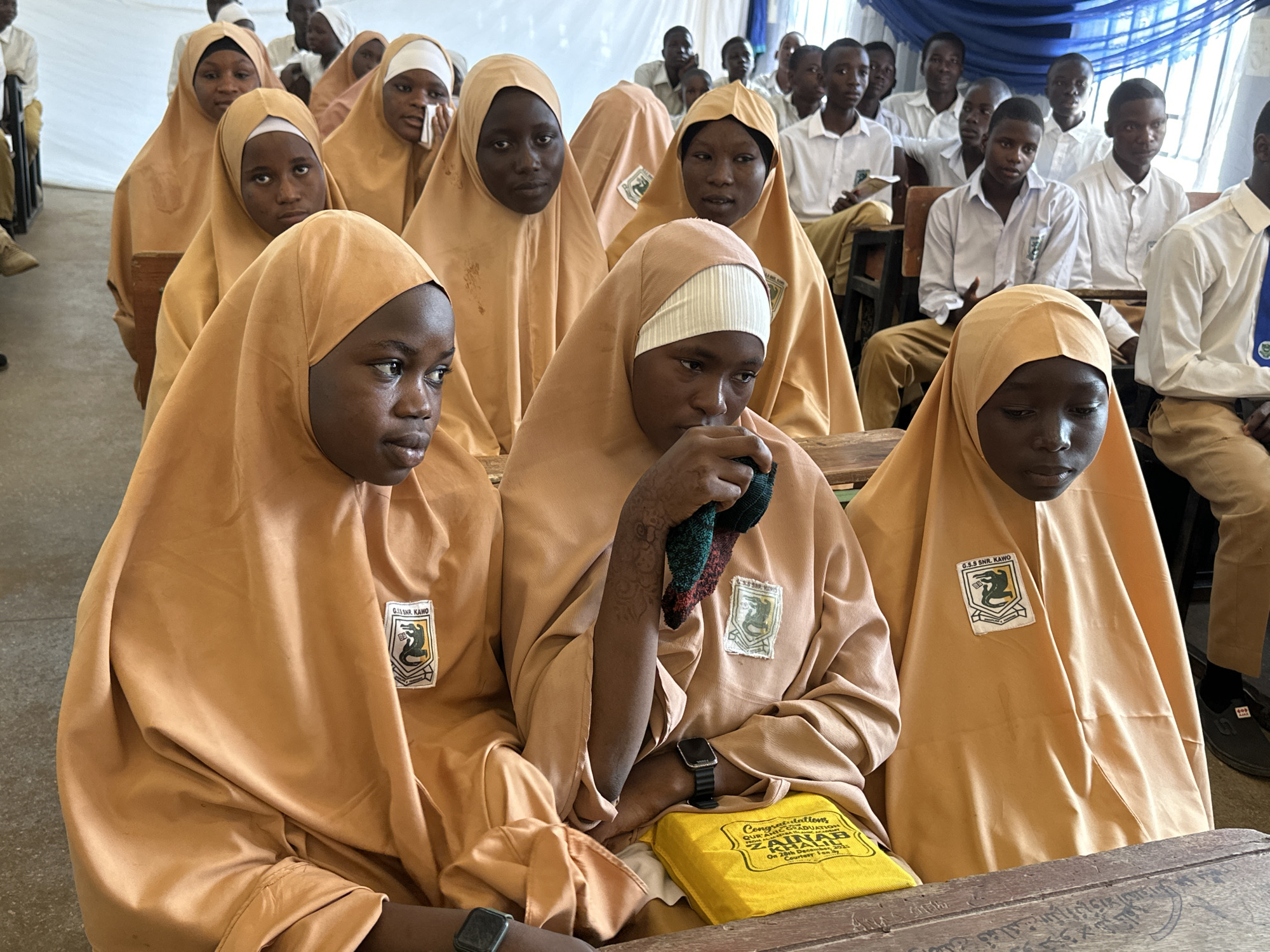 A large group of young students, primarily girls, are seated indoors, likely in a school assembly hall or classroom. In the foreground and mid-ground, several girls are prominently featured, all wearing matching light brown or tan full-body hijabs, with white underscarves visible on some. They are seated at rustic wooden desks or benches. Each girl's garment bears a rectangular patch on the upper left chest, which reads "G.S.S SNR. KAWO" along with an emblem featuring a snake-like creature and a shield.
One girl in the central foreground holds a small, crumpled, multi-colored (green, red, and black) cloth in her hand and wears a black digital watch on her left wrist. On the desk in front of her is a vibrant yellow fabric bag or pouch. Text on this bag is clearly visible and reads: "Congratulations QUR'ANIC GRADUATION ZAINAB KHALIL On 28th May, 2023 Courtesy: For My...". Another girl in the mid-ground is seen wiping her face with a white tissue.
In the background, a group of boys are seated, dressed in white button-up shirts and light-colored trousers, consistent with school uniforms. One boy wears a blue tie. The overall setting is brightly lit, suggesting daytime, with a white fabric backdrop on the left and a blue draped fabric on the right. The students generally appear calm and attentive, some gazing towards an unseen focal point. The visible text on the badges identifies the school as "G.S.S SNR. KAWO," and the message on the yellow bag indicates a Qur'anic graduation ceremony for a student named Zainab Khalil on May 28, 2023, pointing to a celebratory academic event at a Government Senior Secondary School in Kawo, Unguwan Dosa, Nigeria.