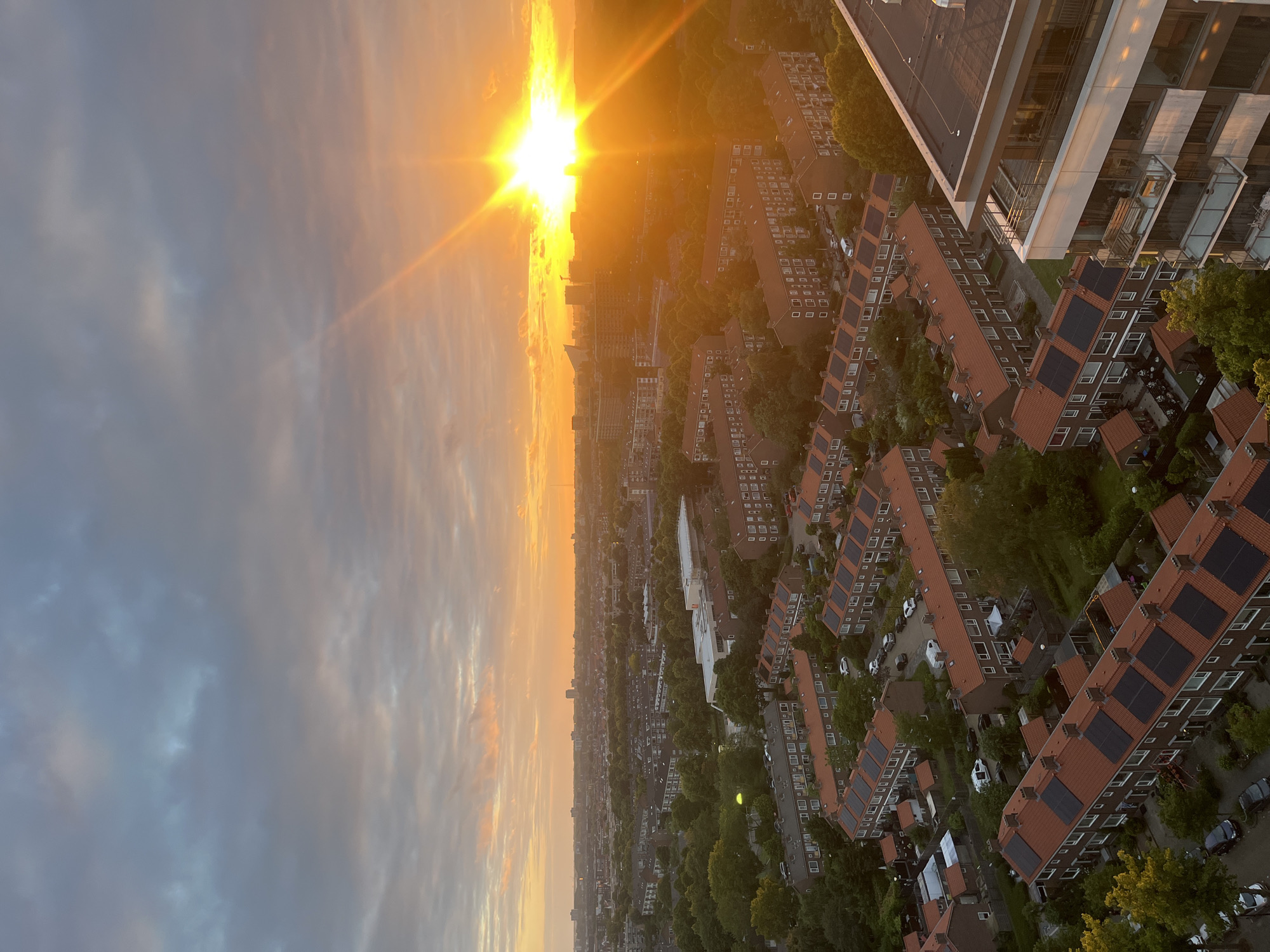 An aerial view captures a cityscape bathed in the warm glow of a sunset. The sky is a gradient of soft oranges and pinks, transitioning into a muted gray at the top, with scattered clouds. A bright, intense sun dominates the right side of the horizon, casting long, distinct rays across the scene. Below, a dense urban environment unfolds, characterized by rows of red-tiled houses with solar panels on their roofs, interspersed with green trees and darker buildings in the distance. Cars are visible on the streets and in driveways, and a modern apartment building with balconies occupies the lower right corner. The overall impression is one of a peaceful evening settling over a residential area.