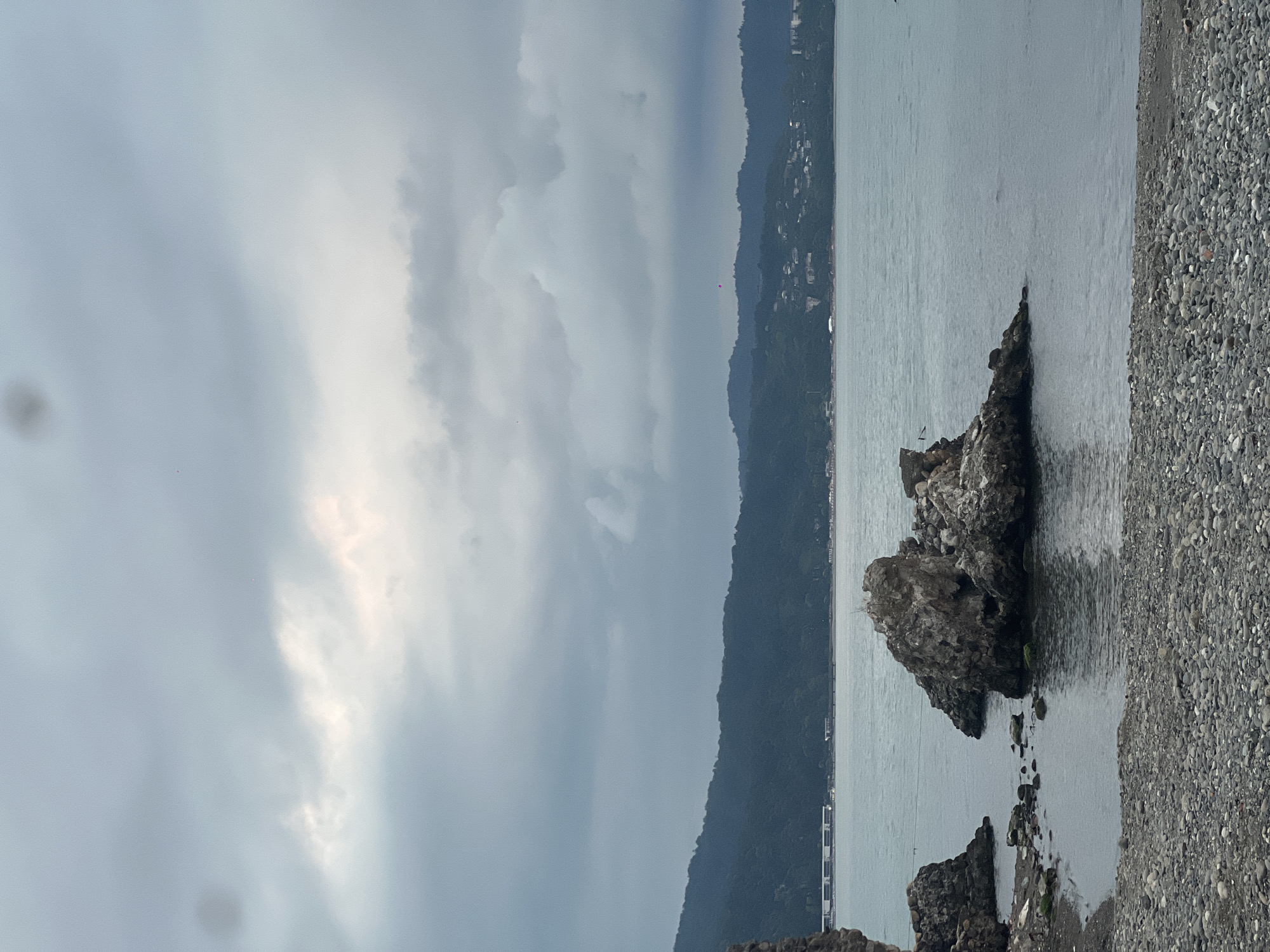 The image depicts a cloudy sky over the Black Sea coast of Sokhumi, Abkhazia. In the foreground, a pebble beach leads to the water's edge. Several large, rocky outcrops rise from the calm sea, some partially submerged. In the middle ground, a line of lush, green hills forms the coastline, dotted with buildings that suggest a developed urban or resort area. The sky is overcast with textured, grey clouds, with a brighter, lighter patch suggesting diffused sunlight. There are no visible people or animals in the frame. The overall atmosphere is serene and slightly somber due to the heavy cloud cover.