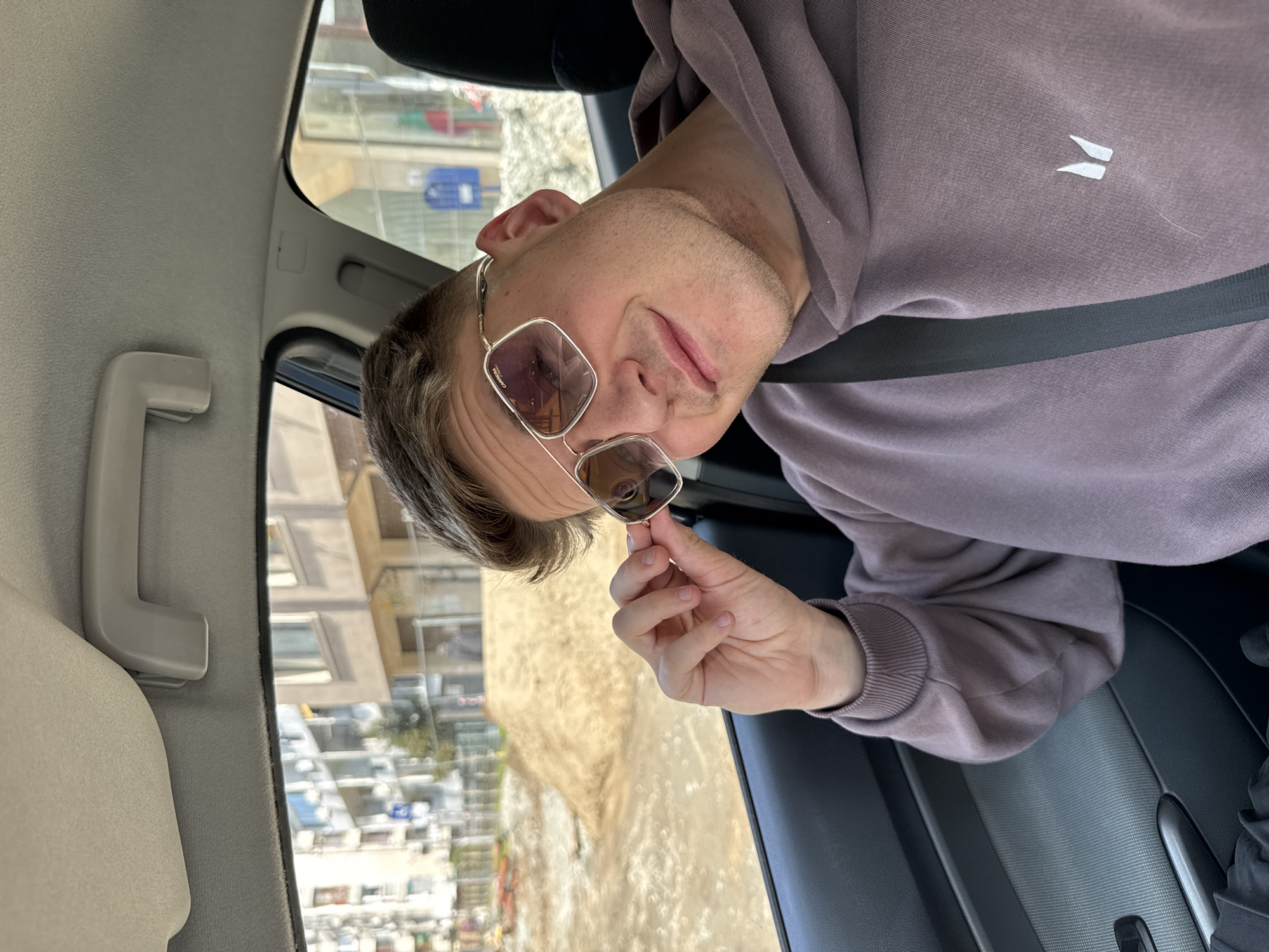 A man wearing a mauve hoodie and silver-framed sunglasses is seated inside a car. He is holding the sunglasses with his right hand, positioning them close to his face. He is wearing a seatbelt. Outside the car window, a street scene in Burgas, Bulgaria is visible, featuring buildings and pedestrian walkways. The interior of the car is light gray and black.