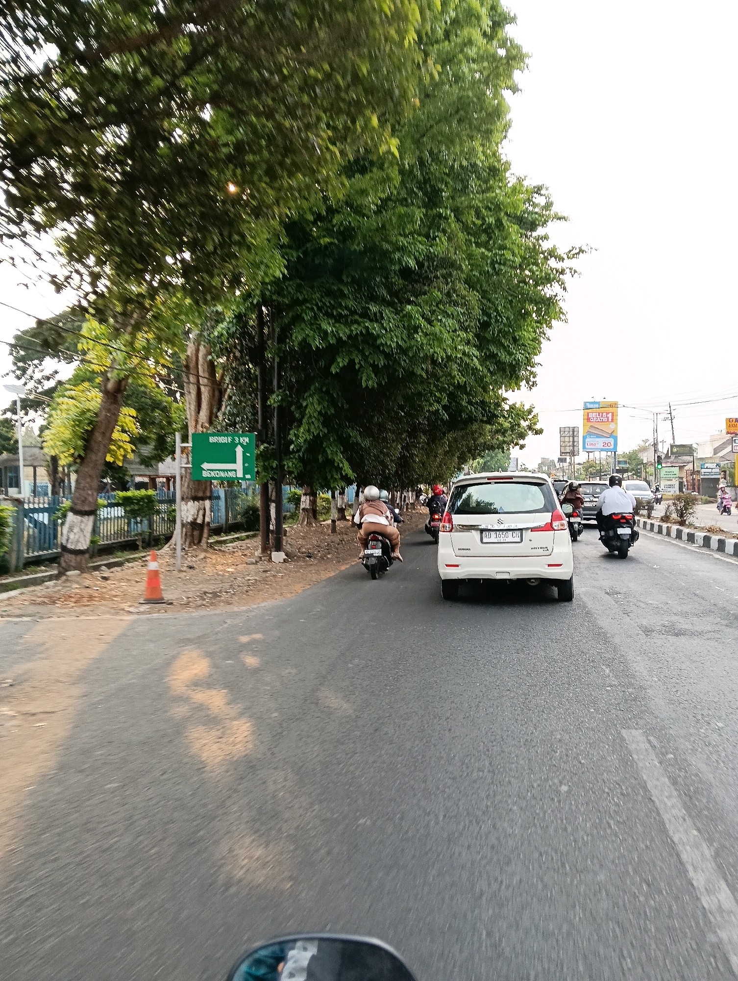 The image depicts a street scene in Jaten, Indonesia, during daylight. The perspective appears to be from a moving vehicle, possibly a motorcycle, looking forward and slightly to the right. The road is asphalt and appears to be a multi-lane street with traffic.
In the foreground, the road surface is visible with some dappled sunlight creating patterns. A side mirror of the vehicle from which the photo is taken is partially visible in the bottom left corner.
Further down the road, several vehicles are present. A white Suzuki Ertiga is in the center of the frame, with its rear facing the camera. Its license plate reads "AD 1650 CH". To the left of the Ertiga, a person on a motorcycle is visible, wearing a light-colored jacket and helmet. Further ahead and to the right, another motorcycle with a rider wearing a white shirt and helmet is moving. Other vehicles, including cars and motorcycles, are scattered throughout the scene, indicating moderate traffic.
On the left side of the road, there is a sidewalk and a grassy area bordered by a blue fence. A traffic sign with arrows and text indicating "BRIGIF 5 KM" and "BERONANG" is prominently displayed on a green pole. An orange traffic cone is placed on the side of the road near the sign. Tall trees with green foliage line the left side of the road, casting shadows.
In the background, buildings and billboards are visible. One billboard displays colorful advertisements. The sky is overcast or bright with hazy sunlight. The overall impression is a typical urban or suburban street in Indonesia during the daytime, with active vehicular traffic.
