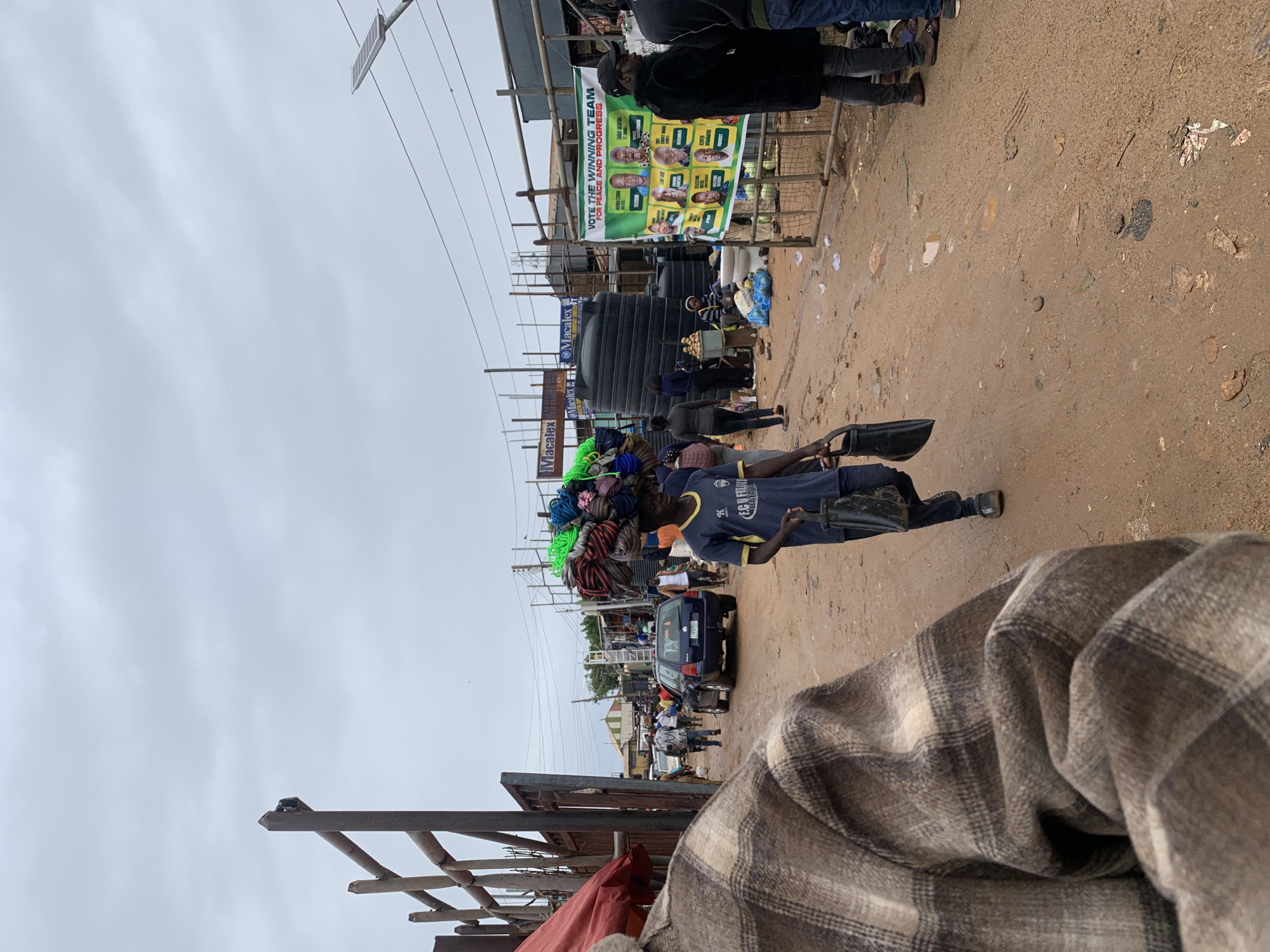 The image captures a street scene in Gura Top, Nigeria, under an overcast sky. The road appears unpaved, with signs of scattered debris. A man in a blue shirt, identified by "F.C UFULINI" and "BATAW" logos, is carrying a load of colorful ropes on his head. In the background, a blue car is visible amidst other pedestrian traffic. Signs for "Macalex" are also visible. There is a political poster displayed, advocating for a "Winning Team" promoting "Peace and Progress" with the names "EMMA AKAFUR" and "QUESTIME" displayed. A black water storage tank is also evident nearby. The lower foreground contains a portion of a checkered flannel garment.
