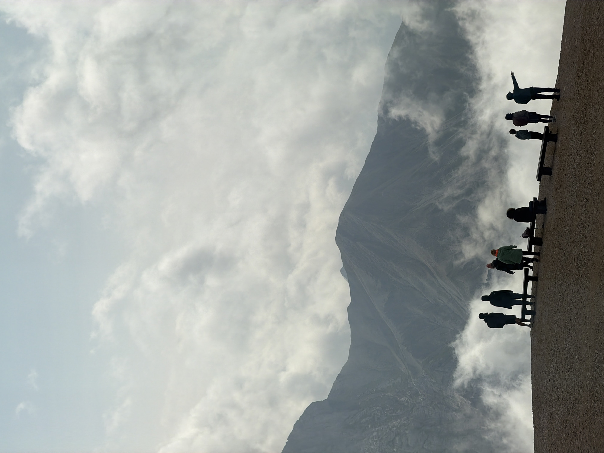 This image depicts a group of eight people, silhouetted against a dramatic mountainous landscape. The people are standing and sitting on a dirt path, some are looking out towards the mountains, and one individual is pointing. There are several benches on the path. The mountains are rugged and imposing, with clouds swirling around them and obscuring parts of the scene. The sky above is overcast. The location appears to be a scenic viewpoint, possibly at a high altitude, given the presence of clouds among the mountain peaks. The overall impression is one of awe and exploration in a grand natural setting.