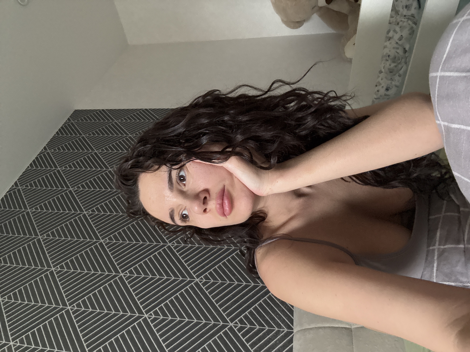 A young woman is captured in a close-up, sideways portrait, lying down in an indoor setting. Her head is turned towards the camera, resting on her right hand, with her fingers gently cupping her chin. She has long, dark, wavy hair spread around her and a neutral, slightly serene expression with her lips lightly parted and dark eyes gazing directly at the viewer. She is wearing a light grey or beige camisole, revealing part of her upper body.
The background features a distinct wall with black and white geometric pattern wallpaper on the left, showcasing intricate intersecting lines forming triangular and square shapes. Adjacent to this, a plain light grey wall is visible. On the right side of the frame, a section of a bed or couch with grey checkered bedding can be seen. Further back, a white piece of furniture, possibly a shelf or headboard, holds a light brown teddy bear, partially obscured. The lighting in the room is soft and even, typical of indoor daytime conditions. No text is visible in the image. The setting is an interior room, located in Chaikovsky, Russia.