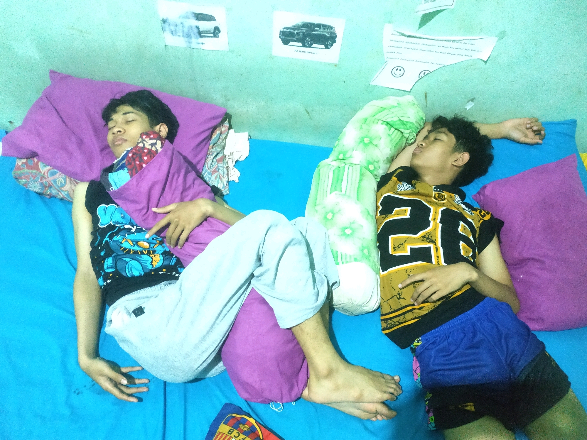 The image depicts two young males sleeping on a blue bed in a room. The room appears to be a living space, possibly a bedroom, with a light green wall. On the wall, there are several pieces of paper attached, including images of cars and text.
The male on the left is lying on his back, propped up by a large purple pillow and a patterned fabric. He is covered partially by a purple blanket and is wearing a black t-shirt with a blue and yellow graphic print and light grey sweatpants. His right arm is bent upwards, resting near his head, and his left arm is extended downwards.
The male on the right is lying on his left side, using a green and white striped pillow. He is wearing a black and yellow sleeveless t-shirt with the number "25" prominently displayed and blue athletic shorts. His right arm is resting across his chest, and his left arm is extended backwards.
Both individuals appear to be asleep, with their eyes closed. The overall environment suggests a casual, domestic setting. The specific location in Pacul, Indonesia, is not visually evident from the image itself, but it is stated as context for the scene.