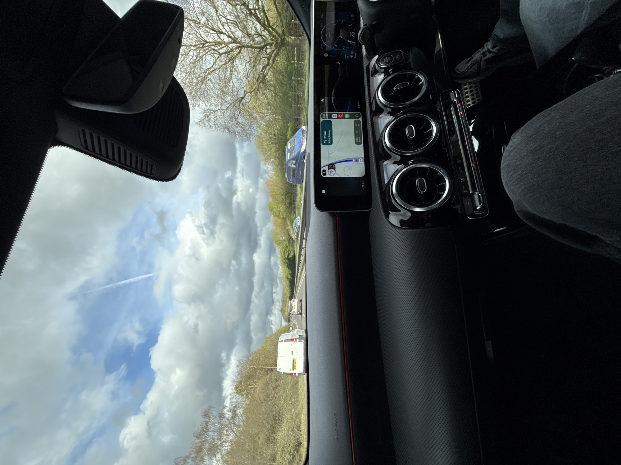 The view from inside a car shows a highway in Mid Suffolk, United Kingdom. The car's dashboard is visible, including air vents and a navigation screen displaying a map. Outside, traffic is present, with vans and cars on the road. The sky is partly cloudy. A contrail is visible. Trees line the highway. The driver is wearing denim jeans and athletic shoes.