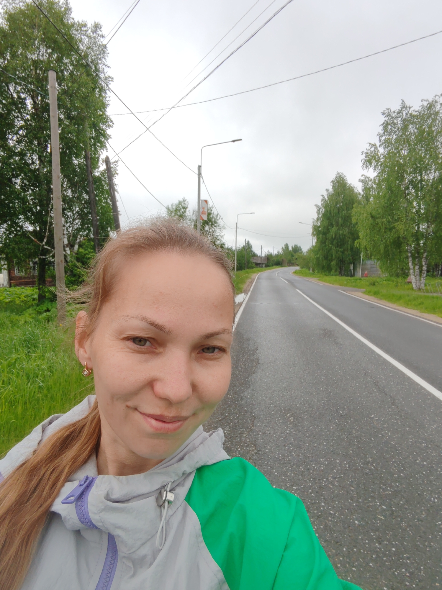 A woman with light brown hair, wearing a green and gray jacket, is taking a selfie on a paved road on a cloudy day. The road, marked with a white center line, curves into the distance, bordered by green grass and trees. In the background, utility poles and streetlights are visible. The location appears to be Чухлэм, Russia, given the surrounding environment and landscape.