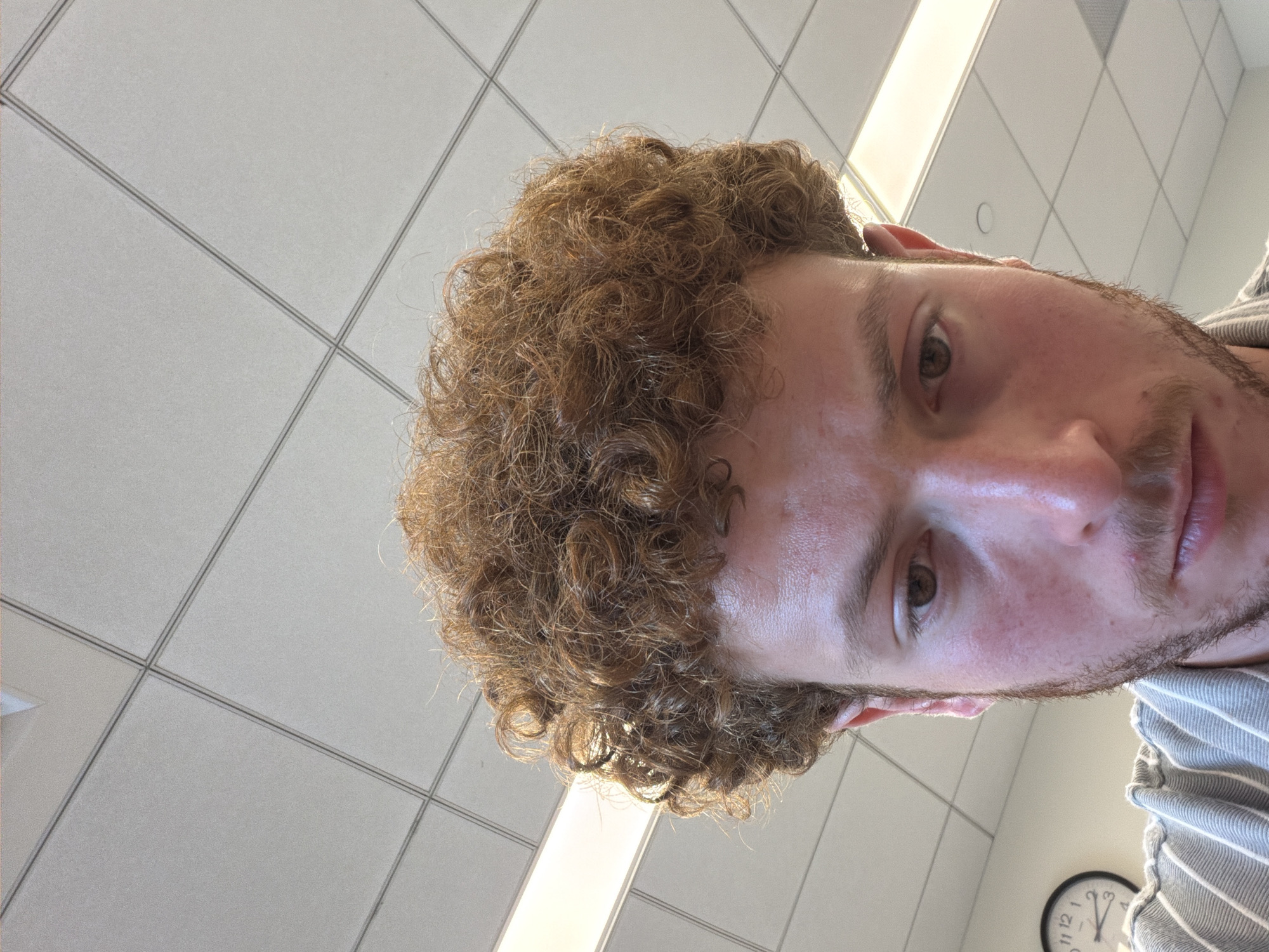 A close-up shot captures a man with curly brown hair and a light beard. He is positioned at an unusual angle, appearing to look directly down at the viewer. His face is well-lit, highlighting his eyes, nose, and lips. The background is an office ceiling with rows of square ceiling tiles and fluorescent lighting. A clock and part of a striped shirt are visible. The location cannot be identified.