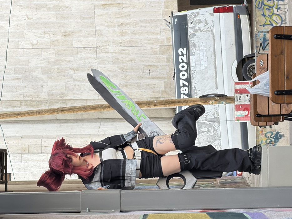 A person is depicted in an urban outdoor setting in Tel-Aviv, Israel, identifiable by Hebrew text "בס"ד" and "7870022" on a white van, and additional Hebrew graffiti on a lower wall. The individual, appearing female, wears a bright reddish-pink wig styled with two prominent upward-jutting sections. Clothing consists of a white strapless crop top, a black and grey cropped jacket, black shorts, a white belt with a yellow strap, black leg coverings with buckles, and black platform shoes. A tattoo is visible on the upper right thigh. The person holds a large prop weapon resembling an oversized pair of grey scissors or a dual-bladed sword with a neon green design on one blade. The individual leans against a grey vertical column with their right foot on the ground and their left foot resting on the prop weapon. In the background, a light-colored, tiled building is visible. A weathered wooden pole stands to the right of the person. Below, a low concrete wall covered in multi-colored graffiti and two wooden benches, one holding a white plastic bag, are in the foreground. A white cargo van with obscured windows is parked behind the pole. A thin utility wire crosses the upper portion of the frame.