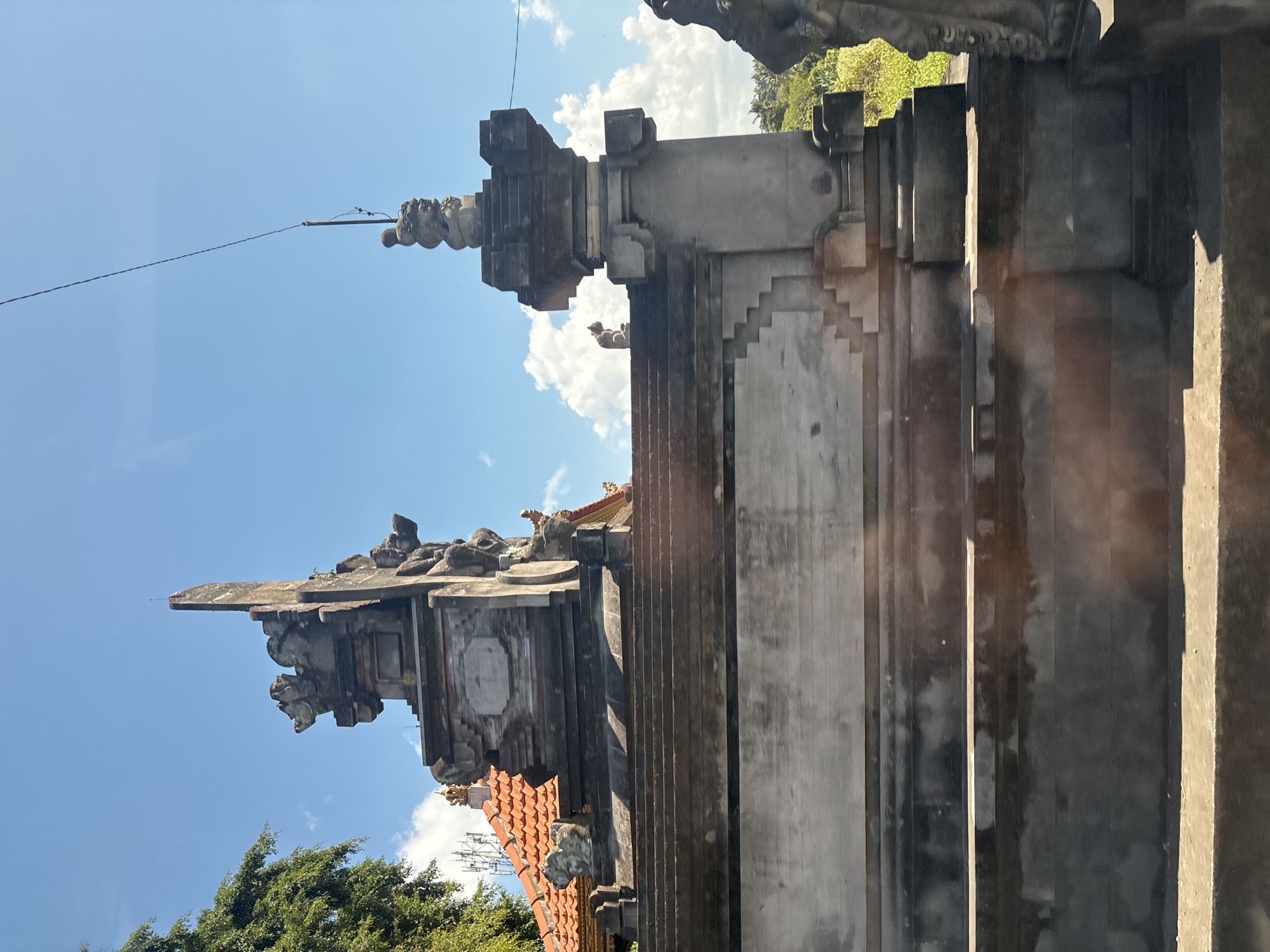 The image shows a traditional Balinese stone structure, possibly a temple gate, in Sukawati, Indonesia. The structure is made of weathered gray stone and features intricate carvings of mythological figures and architectural elements. A steep staircase leads up to the structure, with several levels and platforms. The sky is bright blue with scattered white clouds. To the left, a large green tree with dense foliage is visible, and in the background, a building with a red tiled roof and communication towers can be seen. The right side of the image shows a grassy area with some trees. The overall scene conveys a sense of ancient architecture and a tropical environment.