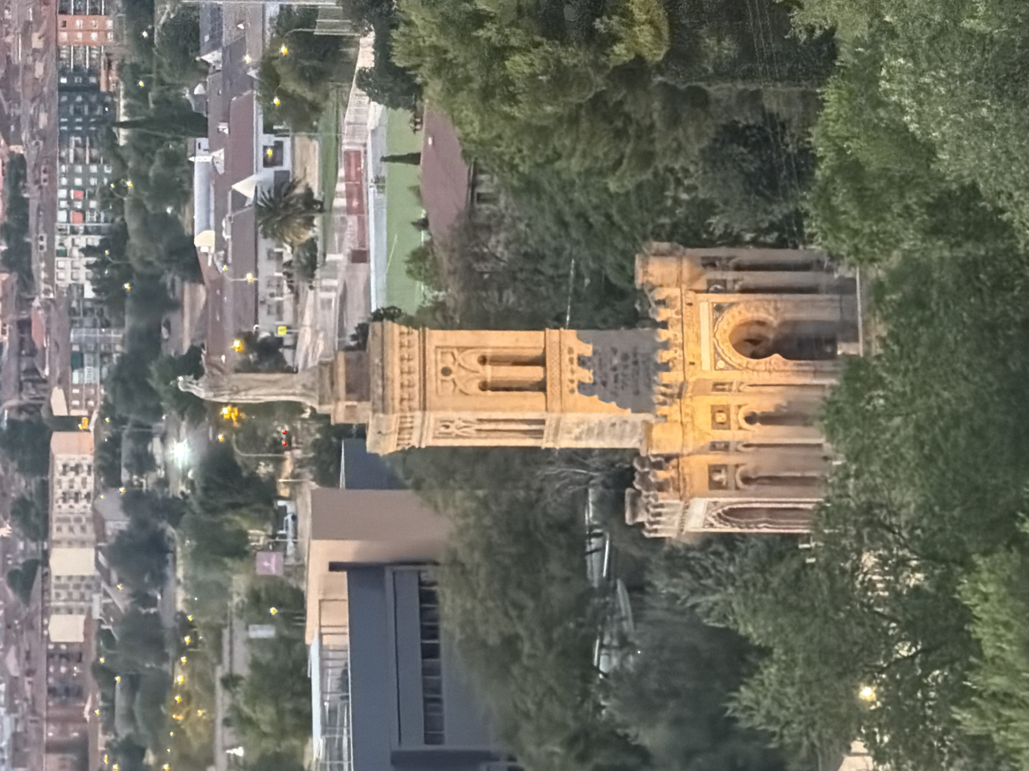 The image, taken from a high vantage point, shows a cityscape of Toledo, Spain, at dusk. A prominent monument, likely the Monumento a la Batalla de Lepanto, stands in the mid-ground, illuminated by artificial lighting, casting a warm glow on its detailed architecture. The monument features a statue of a standing figure at its apex and is surrounded by crenellated battlements and ornate arches.
The monument is nestled among lush green trees, with the dense foliage partially obscuring the lower levels of the structure. Behind the monument, a stadium with a green field and red running tracks is visible, bordered by a white fence. Further in the background, a residential area with multi-story buildings, some with red-tiled roofs, stretches across the horizon. The sky is a dusky grey, with scattered lights twinkling from streetlights and windows in the city. The overall scene conveys a sense of historical grandeur against a backdrop of modern urban life.