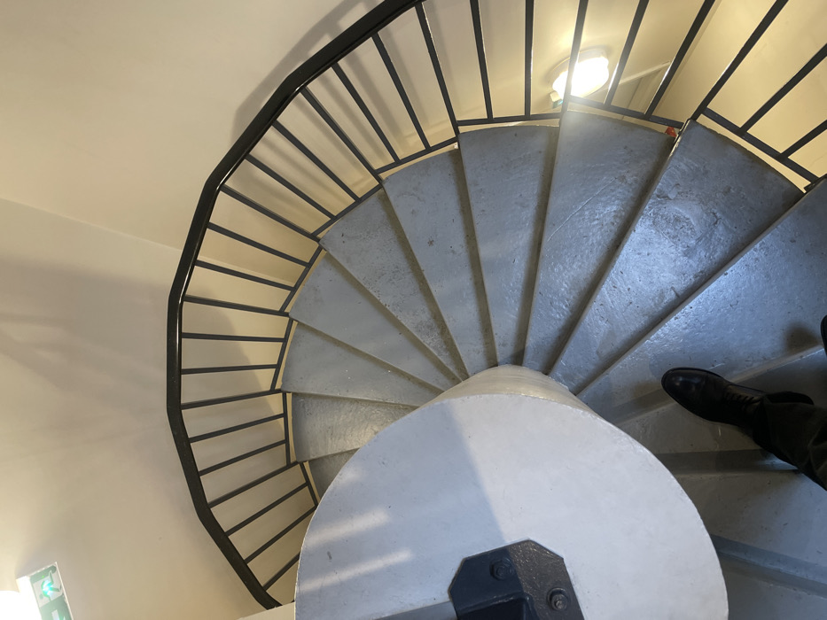 The image displays a high-angle view looking down a concrete spiral staircase inside a building in Montigny-le-Bretonneux, France. The staircase features light grey, wedge-shaped treads spiraling clockwise around a substantial, circular central newel post painted white. A dark, possibly black, metal balustrade with multiple vertical bars extends along the outer edge of the steps. The surrounding walls are painted a light cream or beige color. Artificial illumination is present, with a circular, frosted light fixture visible on the lower right wall. An emergency exit sign, green with a white running man pictogram and arrow, is mounted on the upper left wall. A person's lower legs and feet, clad in dark, polished leather boots, are positioned on one of the lower steps, indicating a descent.