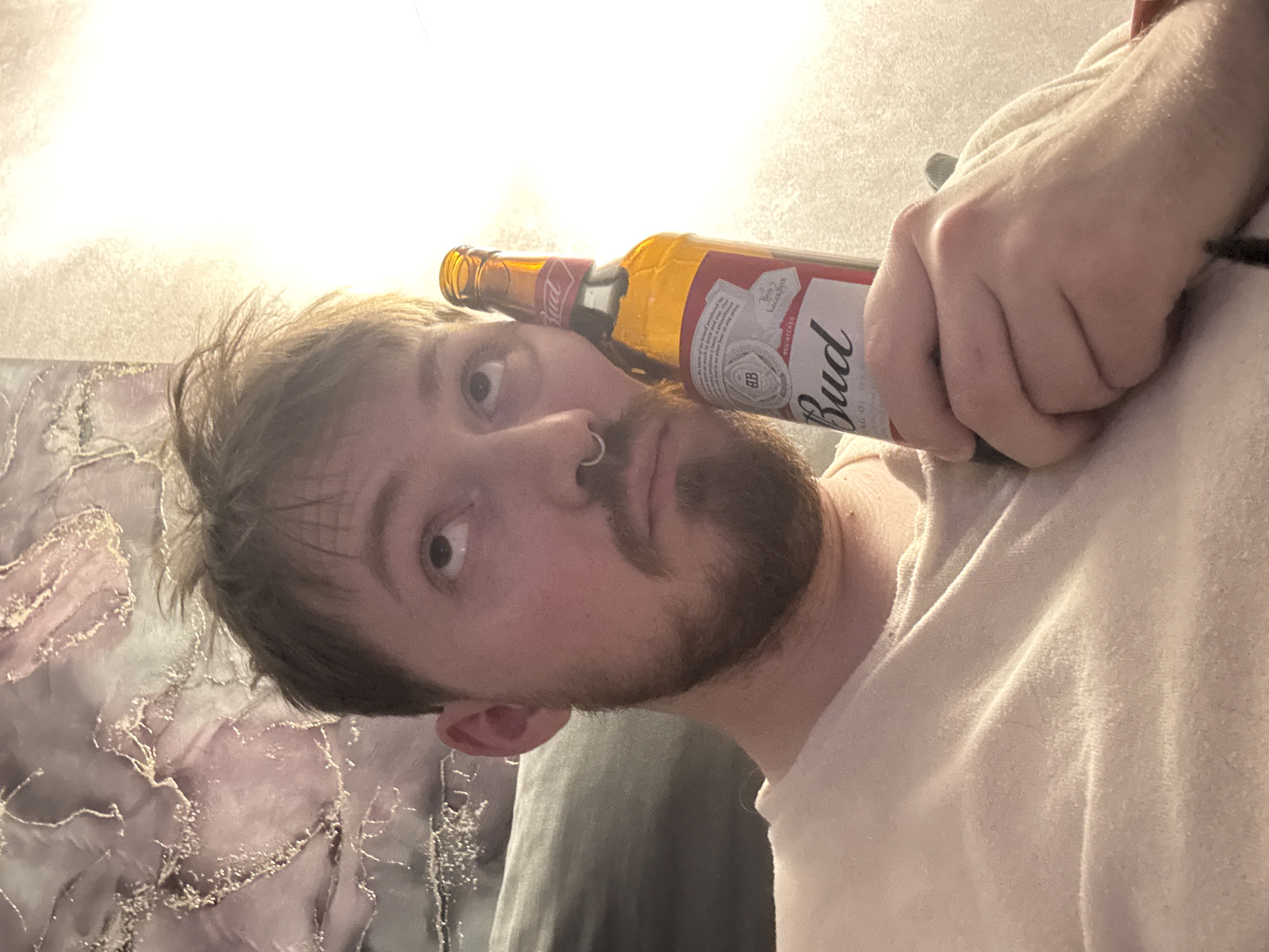 A close-up, tilted perspective shot shows a light-skinned man with light brown hair and a beard. He is holding a bottle of Budweiser beer against his head, near his ear. He has a nose ring. He is wearing a light-colored t-shirt. The background includes a wall and a patterned object that could be a painting. It is not possible to determine the location as Chaikovsky, Russia from the provided image.