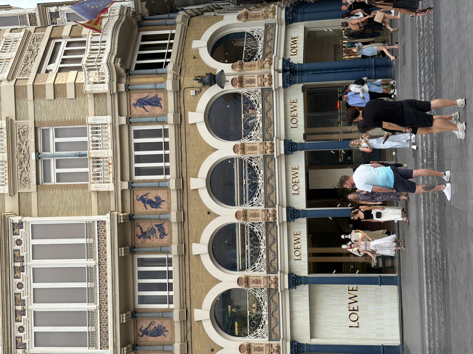 The image shows the exterior of a LOEWE store on a city street. The building has an ornate facade with multiple levels, featuring arched windows, decorative tiles with figures, and balconies. The LOEWE logo is visible on the store's front. Several people are standing and walking on the sidewalk in front of the store, some of whom appear to be shoppers or passersby. The street in front of the building is paved. The overall scene suggests a commercial area in a city.