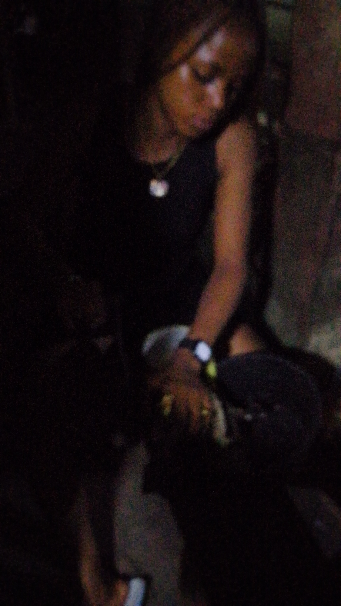 A person wearing a dark top and a necklace is looking down. They are also wearing a watch on their left wrist. The scene appears to be dimly lit.