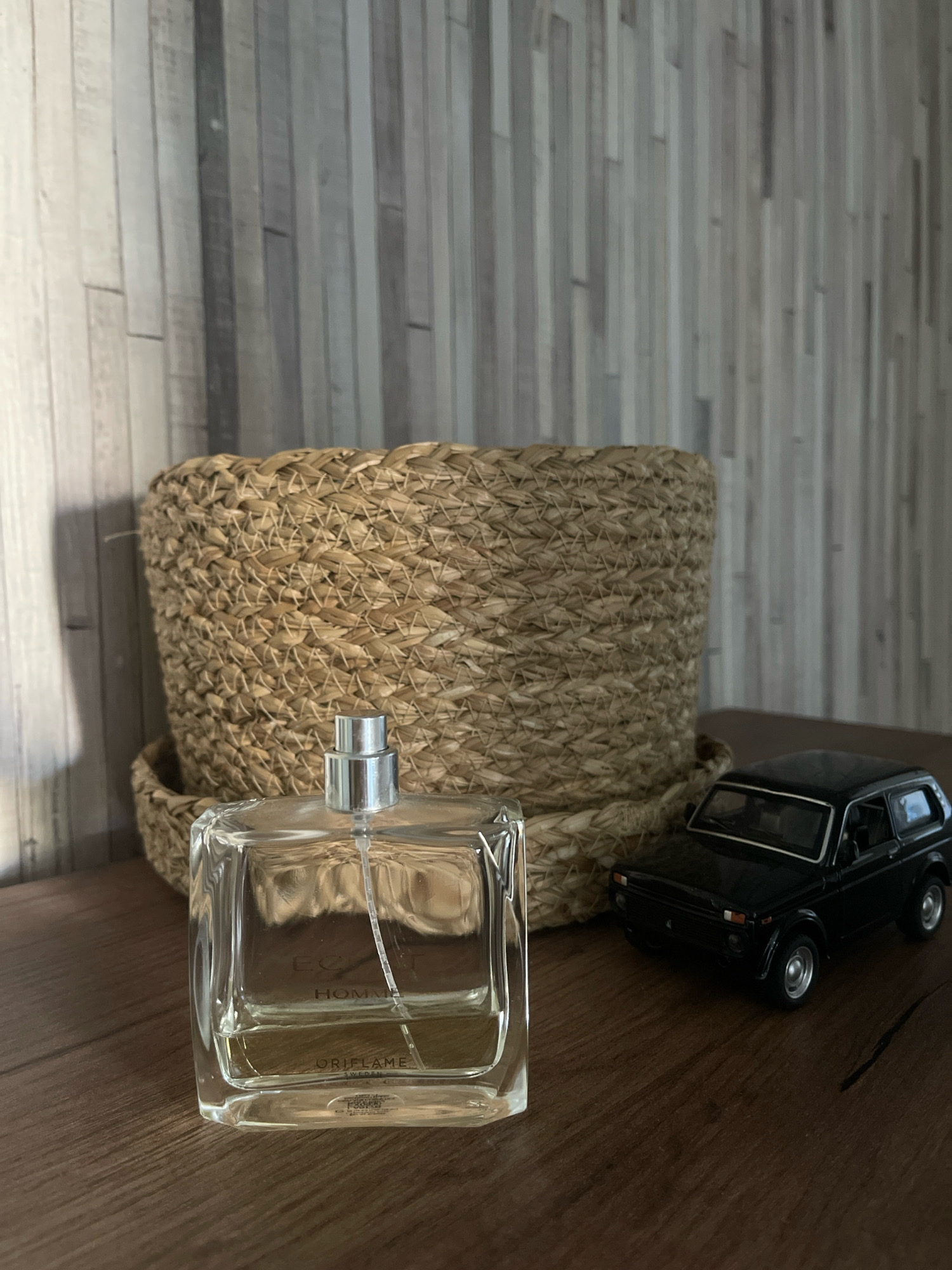 The image displays a still life composition on a wooden surface. In the foreground, a clear glass perfume bottle with a silver cap sits prominently. The bottle contains a light-colored liquid and features branding that includes "EC" "HOMME" and "ORIFLAME SWEDEN". Behind the perfume bottle, a large, woven straw basket is positioned. To the right of the basket, a black toy car, resembling a vintage SUV, is placed. The background consists of a wall with vertical wooden planks, varying in color from light gray to dark gray. The lighting suggests an indoor setting, casting shadows from the objects onto the wooden surface and the wall. There are no people or animals present in the image. The location in the city of Tallinn, Estonia, is not discernible from the visual information provided in the image.