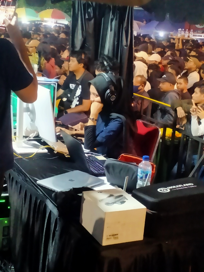 The image captures a scene from a crowded outdoor event in Boyolali, Indonesia. The scene is taken from a low angle, and the subject is a young woman wearing a blue hijab, sitting at a table with a laptop and a microphone. She is wearing headphones and is looking at a screen. She is working on a laptop, there is a water bottle, a black case, and a white box on the table. To her left, a man in a black T-shirt is also working on a laptop. Behind her, the image shows many people standing and sitting in rows. The scene appears to be an event with a stage, and the crowd is watching a performance. The event is taking place in the evening, as evidenced by the dim lighting and shadows. There are several other objects in the image, including a computer monitor, a speaker, and a few chairs. The environment is characterized by large black fabric, a black table, and a large crowd of people. The scene is likely taking place at an outdoor event, such as a concert or festival.