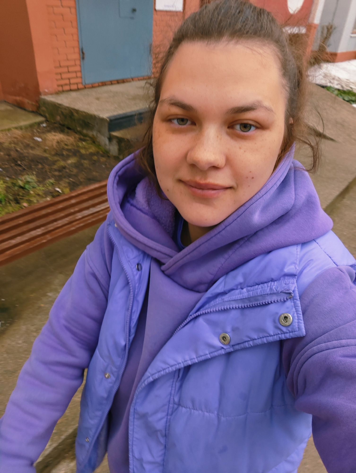 A woman with brown hair is wearing a light purple hooded sweatshirt and a light purple puffy vest. She is standing outside in front of a building with a red brick wall and a blue door. There is a bench to her left and snow on the ground. The location is Chaikovsky, Russia.