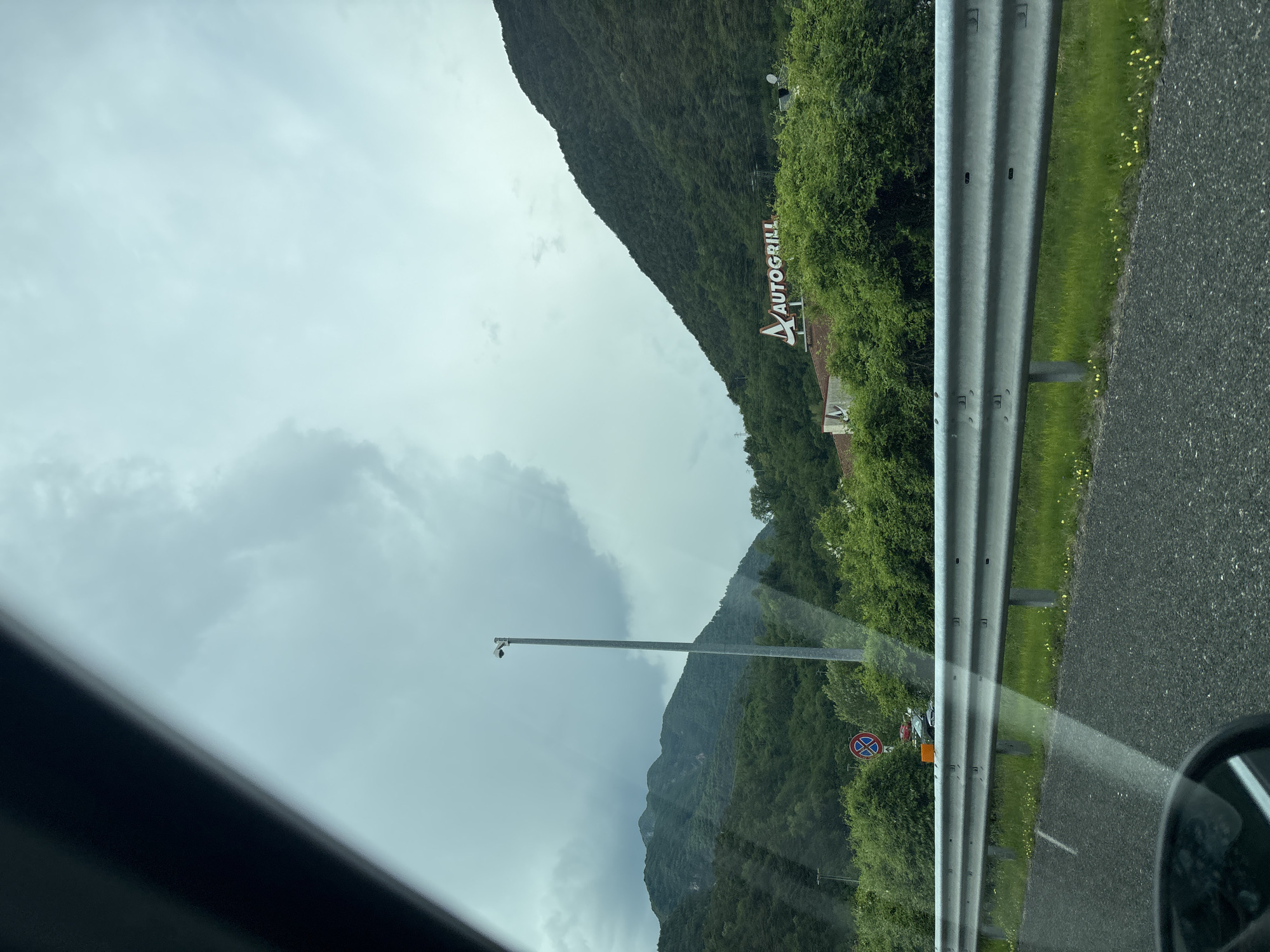 The image, taken from inside a vehicle, shows a roadside scene in Moggio Udinese / Mueç, Italy. Visible outside the car window is a highway with a guardrail. Beyond the guardrail, there's a green, mountainous landscape. A sign for "Autogrill" is visible, indicating a rest area or restaurant along the highway. A tall light pole stands near a 'no parking' sign. The sky is overcast.