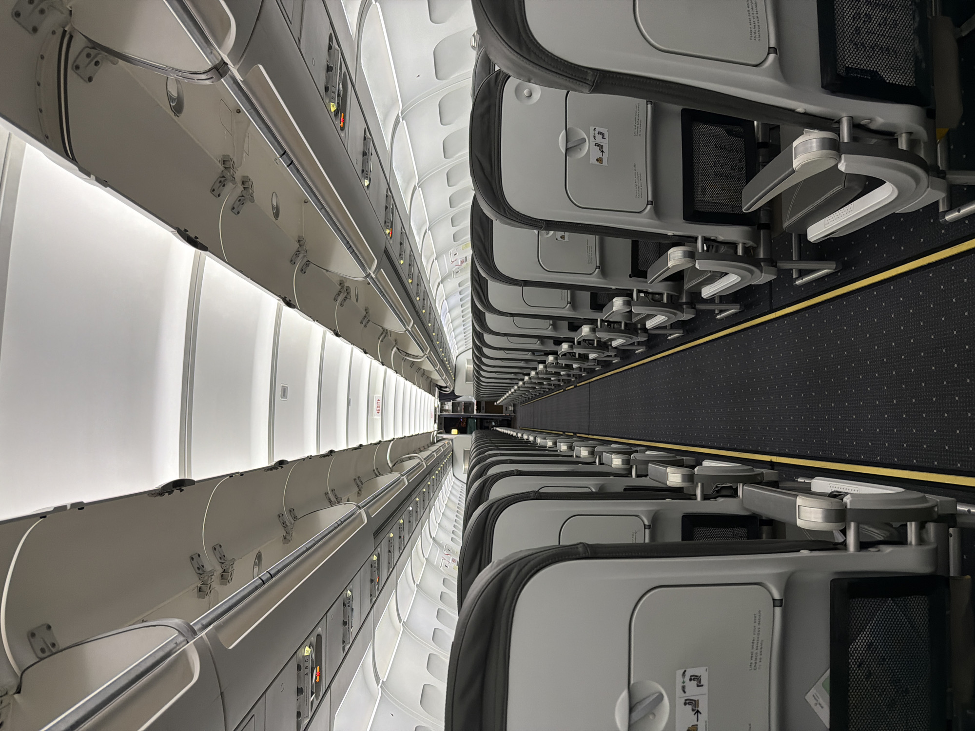 This is an interior shot of an airplane cabin, most likely taken during boarding or deplaning, as no passengers are visible. Rows of seats line both sides of a central aisle carpeted with a dark-patterned design, featuring a yellow stripe on either side. The seats are predominantly gray with dark headrests, and each has a tray table in the upright position. Overhead compartments run along the length of the cabin, with reading lights and air vents visible above each row of seats. The overall lighting is bright, creating a clean and sterile environment. The image is taken from a slightly elevated perspective, looking down the length of the airplane. There is nothing indicating the image was taken in Communauté rurale de Diass, Senegal.