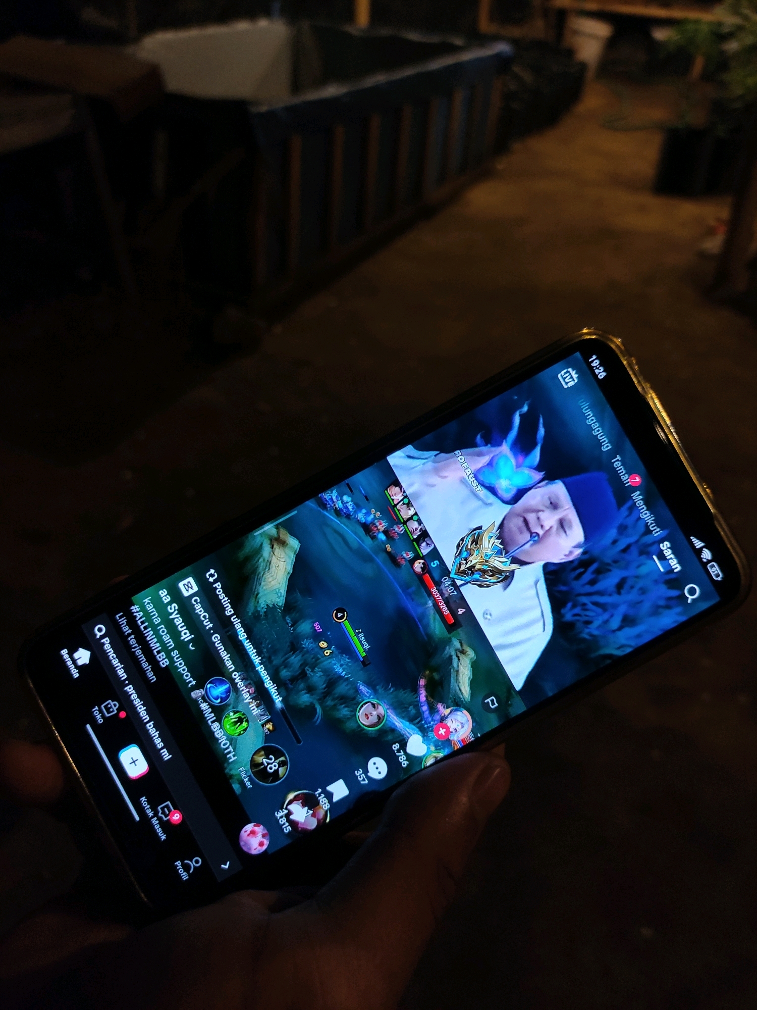 The image shows a person holding a smartphone, displaying a live streaming video, likely from the app TikTok. The video features an elderly man in a white shirt and blue headwear, with a blue ethereal glow emanating from his hands. The phone screen also shows various interface elements typical of a live stream, including user icons, comments, and engagement metrics like likes and viewer counts. The hashtag #ALLINMLBB is visible, suggesting the stream is related to the mobile game Mobile Legends: Bang Bang. The background is dark and out of focus, but appears to be an outdoor setting at night, possibly a backyard or patio area. The presence of what looks like a dark, weathered wooden structure and some foliage suggests a residential or semi-rural environment. The time displayed on the phone is 19:26. The location is identified as Tiudan, Indonesia.