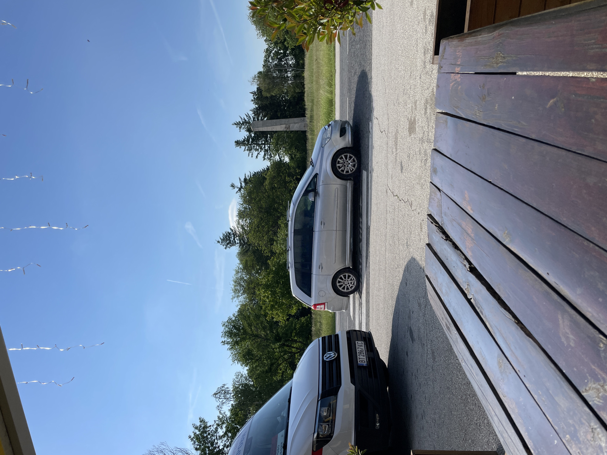 The scene is in Sheynovo, Bulgaria. Two vehicles, a silver minivan and a white van, are parked on a paved road. A wooden bench is adjacent to the road. The sky is blue with streamers or ribbons hanging in the air. Trees are visible in the background.