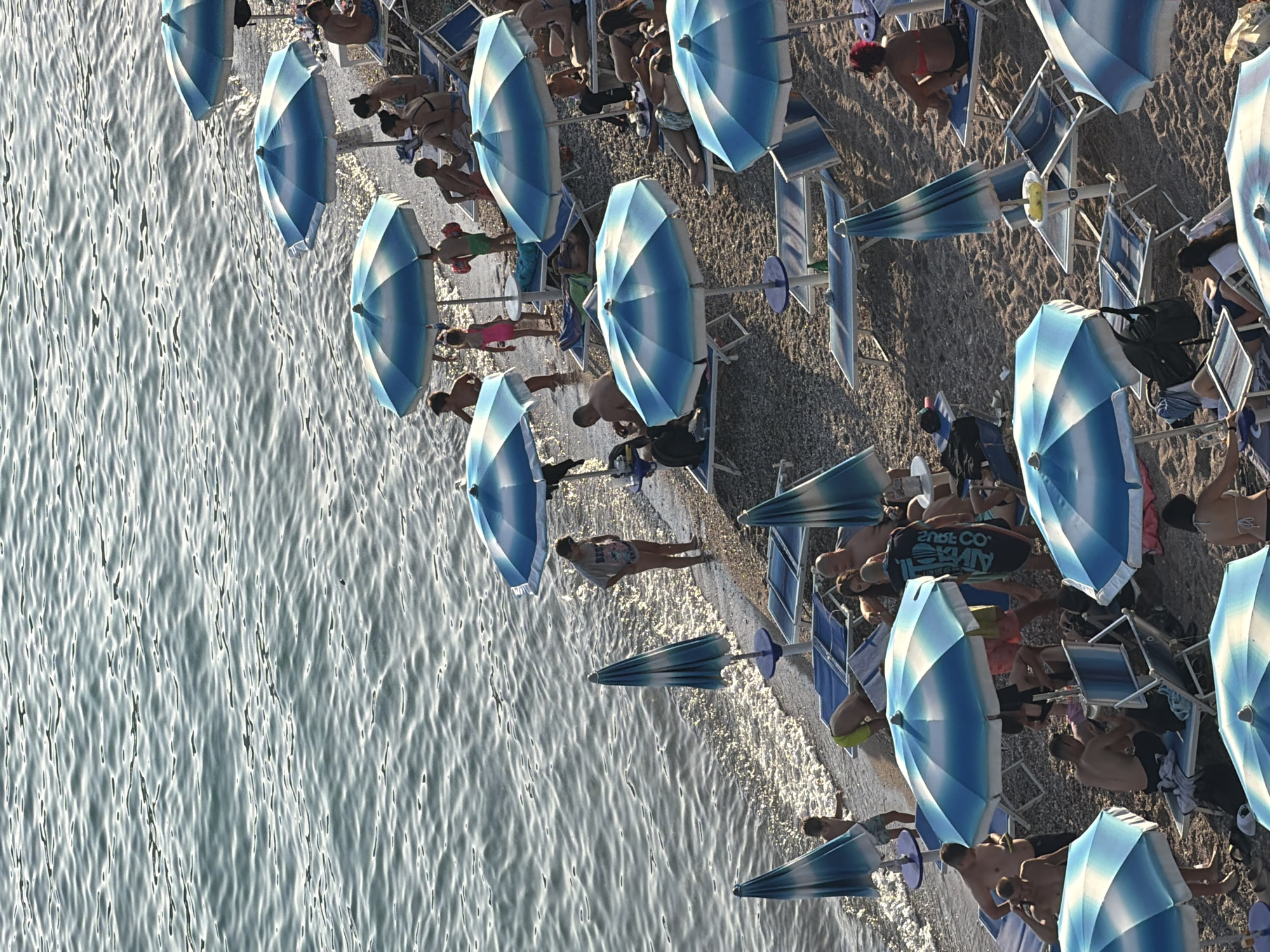The image depicts a crowded beach in Boreti, Montenegro. The scene is dominated by a body of water on the left and a sandy beach on the right, occupied by numerous beachgoers. Several blue and white striped umbrellas are scattered across the sand, providing shade. People are seen lounging on beach chairs, walking along the shore, and standing in the water. Some individuals are partially submerged in the water, while others are sunbathing or sitting under umbrellas. The water appears calm with gentle ripples. The overall atmosphere is one of a busy summer day at the beach.