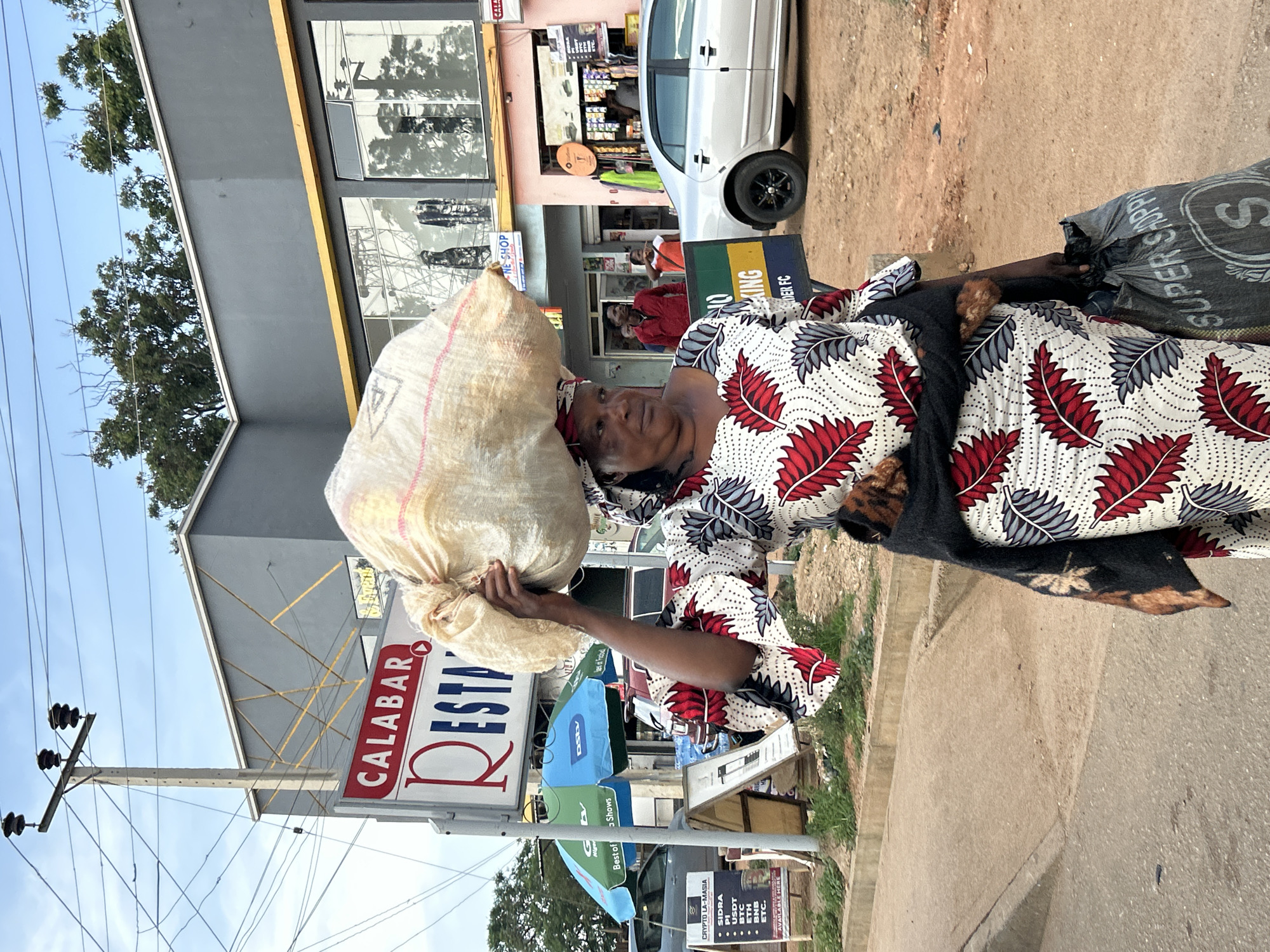 A woman in Jos, Nigeria, walks along a street carrying a large sack of goods on her head and a shopping bag in her right hand. She wears a traditional patterned dress and a headwrap. Behind her, a restaurant sign reads "CALABAR RESTAURANT." Several shops and a silver car are also visible. The sky is cloudy, and trees line the background.