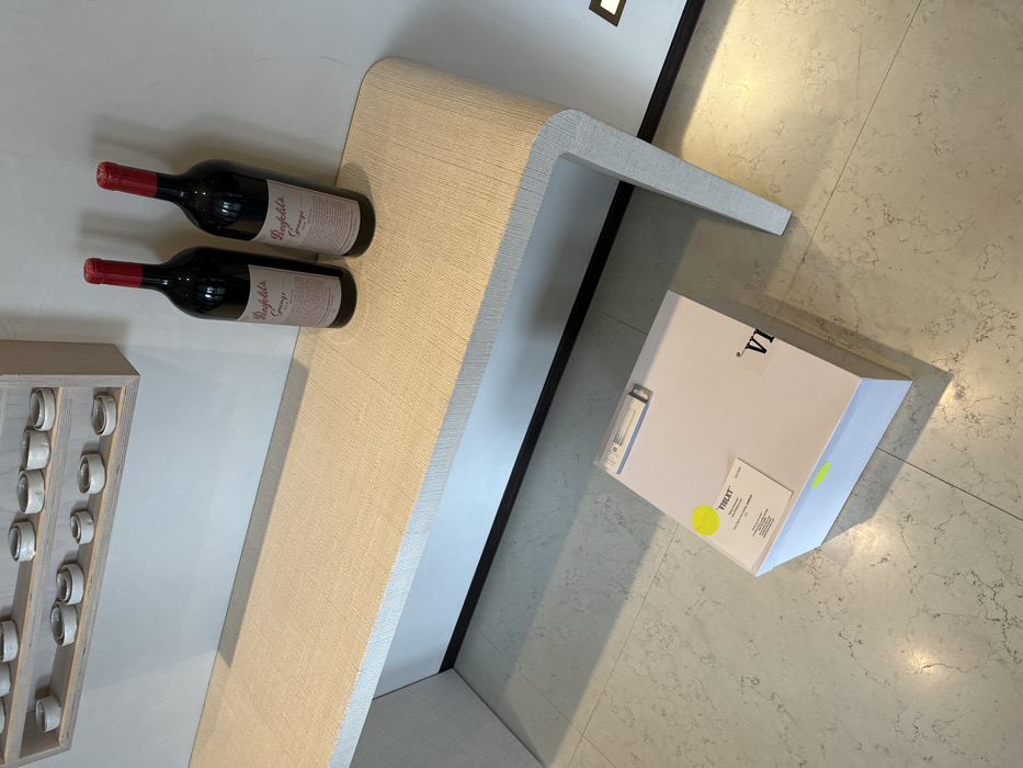 Two bottles of Penfolds Grange wine lay on a beige, textured console table. To the left is a framed display with multiple rows of small, white, ring-shaped objects. To the right of the table is a white box with a yellow sticker, resting on a marble floor. The composition is shot from an overhead angle, highlighting the objects' arrangement on and around the console table.