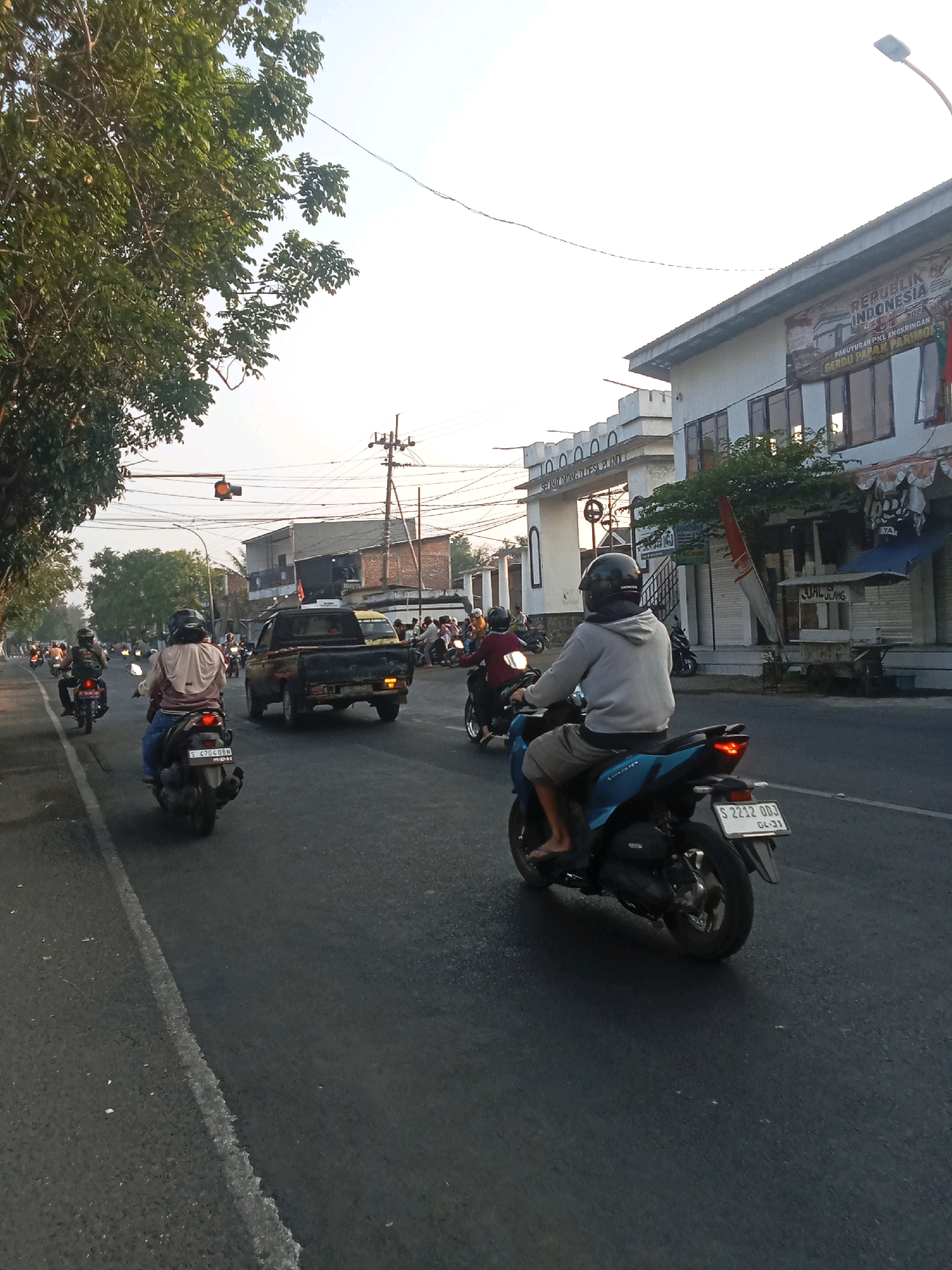 The image captures a street scene in Kaliwungu, Indonesia, likely during the late afternoon or early morning, given the low angle of sunlight and long shadows. The foreground is dominated by a paved road with a white dashed line marking the lane. Several motorcycles are present, with a blue scooter in the immediate foreground, its rider wearing a grey hoodie and shorts, and a black helmet. Another scooter is visible to the left, with a rider in a light-colored jacket and blue jeans, also wearing a helmet. Further down the road, a black pickup truck is traveling in the same direction, with traffic lights visible overhead. On the right side of the road, a white building with multiple windows and awnings is situated. A banner with text in Indonesian, including "REPUBLIK INDONESIA," is attached to the building. Trees line the left side of the road, their branches extending towards the sky. The overall environment suggests a busy urban or semi-urban street with moderate vehicular traffic.