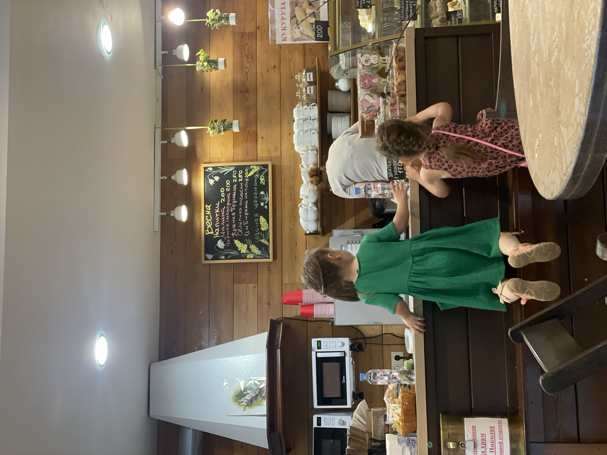 The image captures an indoor scene, likely a café or bakery, situated in what appears to be Khimki, Russia. Two young girls, dressed in a green dress and a pink patterned dress respectively, stand at the counter where an adult, presumably a cashier or staff member, is tending to them. The girls seem to be looking at the items displayed on the counter.
Behind the counter is a wooden wall adorned with hanging plant decorations and a chalkboard menu written in Russian. The menu advertises various drinks and pastries, including those with names such as "Malina" and "Black Currant".
There is also a glass display case filled with baked goods, hinting that the café offers a variety of sweets. In the foreground, a table with a round, light-colored surface is partially visible, along with a wooden chair. The interior is lit with recessed ceiling lights and decorative lights along the wooden wall. Near the girl in the green dress are a stack of red cups and a microwave. Bottles of water and various other items are visible near the bottom of the image.