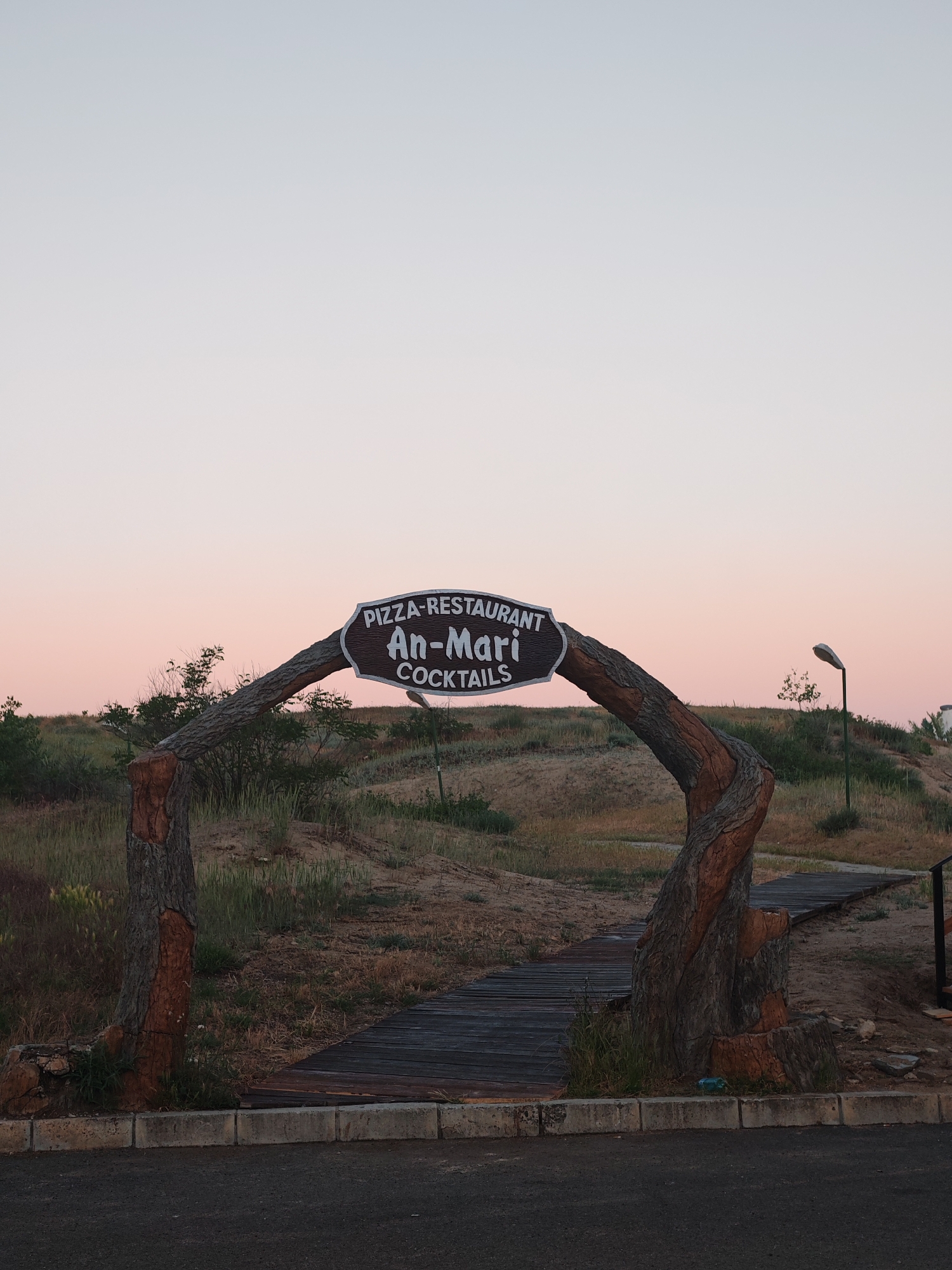 Captured at dawn in Nesebar, Bulgaria, the image showcases the entrance to "An-Mari," a Pizza-Restaurant serving Cocktails. The entrance is defined by a rustic arch crafted from what appears to be modified tree trunks, supporting an oval sign displaying the establishment's name and offerings. A wooden boardwalk extends from the archway, leading towards a grassy, sandy incline dotted with sparse vegetation. A solitary street lamp stands to the right of the path. The foreground reveals a dark asphalt surface with a concrete curb, marking the immediate approach to the entrance.