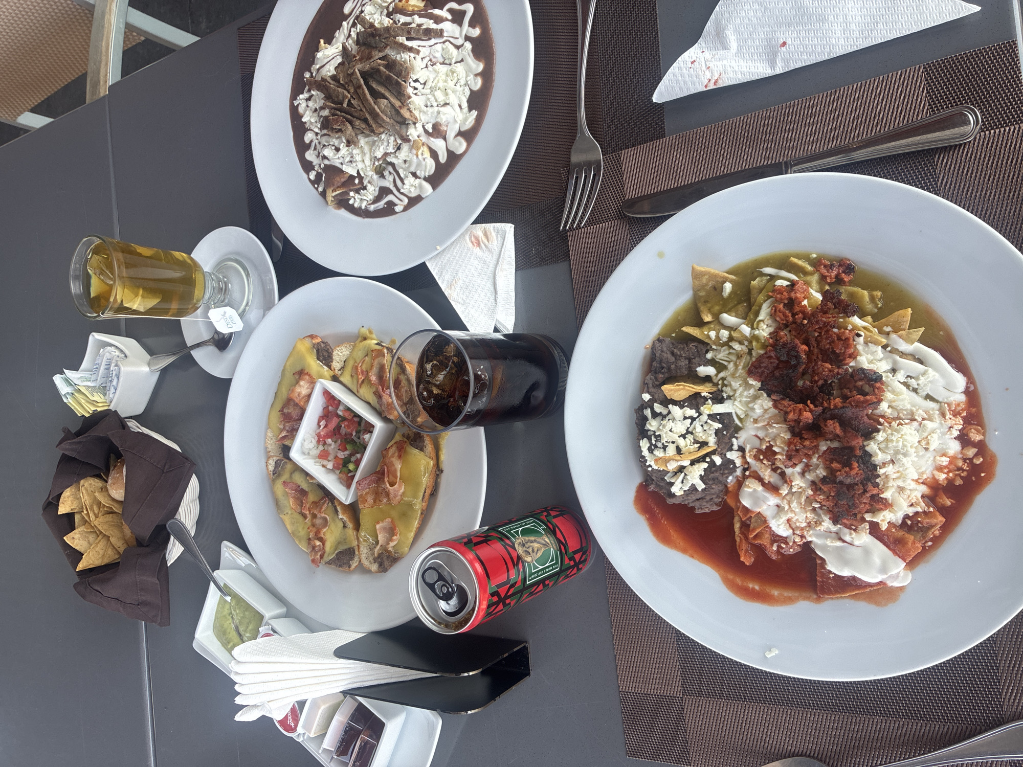The image shows a tabletop scene with various dishes and beverages, likely in Pueblo Nuevo de Jasso, Mexico. Three plates of food are visible: one with enchiladas covered in mole sauce and sprinkled with cheese and onions, another with enchiladas possibly filled with meat or cheese and topped with a small container of pico de gallo, and a third with chilaquiles (tortilla chips simmered in salsa) accompanied by beans, chorizo, and sour cream or cheese. A basket of tortilla chips with containers of guacamole is present. Beverage items include a glass of iced tea, a glass of cola, and a can of soft drink. Cutlery, napkins, sugar packets, and condiments are arranged around the dishes. The table surface is dark gray, with a brown textured placemat on the right. The overall setting suggests a meal at a restaurant or outdoor dining area.