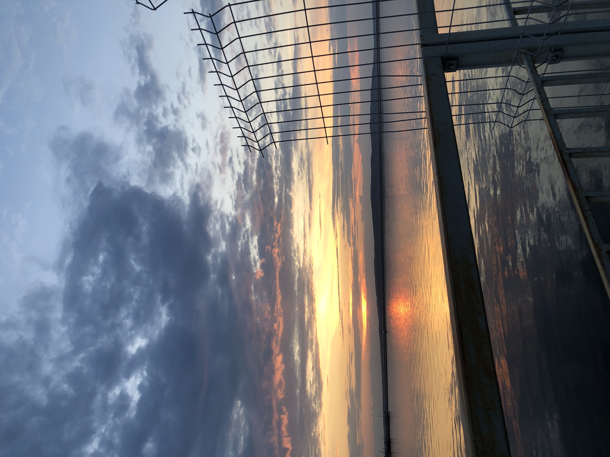 The image depicts a sunset viewed through a wire fence and a metal structure with a ladder. The sky displays a gradient of colors, with dark clouds transitioning to orange and yellow hues near the horizon. The setting sun reflects on the water, creating a bright, shimmering path. The location is likely Chaikovsky, Russia, given the request. The scene is devoid of people. The primary objects are the fence, the metal structure with the ladder, the sky, and the water.