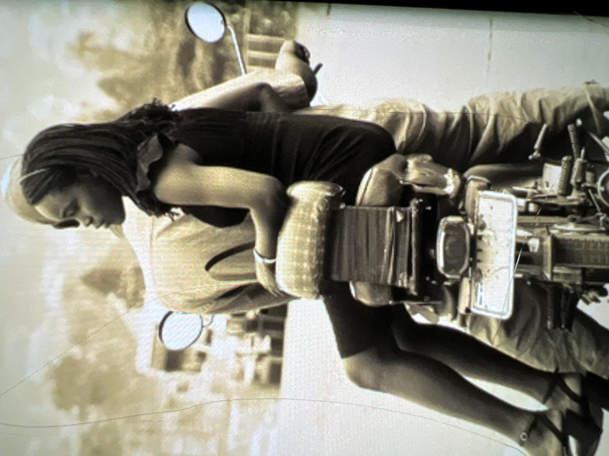 The image depicts a young woman riding on the back of a motorcycle, driven by a man whose upper body is largely obscured by the woman. The woman is wearing a dark, possibly black, sleeveless dress that reaches her mid-thighs, and dark stockings or tights. She has high-heeled sandals on her feet. Her hair is dark and styled in braids or a similar fashion, with a thin band across her forehead. She is looking to her left, away from the camera. The man driving the motorcycle is wearing a light-colored, possibly khaki, shirt and trousers. The motorcycle itself has a passenger seat with a textured, cushioned backrest. A license plate is visible at the rear of the motorcycle. The background appears to be an outdoor urban environment, with blurred buildings and possibly trees. The overall impression is of a candid street scene. The image's quality suggests it might be a scan of an older photograph, with some visible scratches or lines on the surface. The location is identified as Lagos, Nigeria.