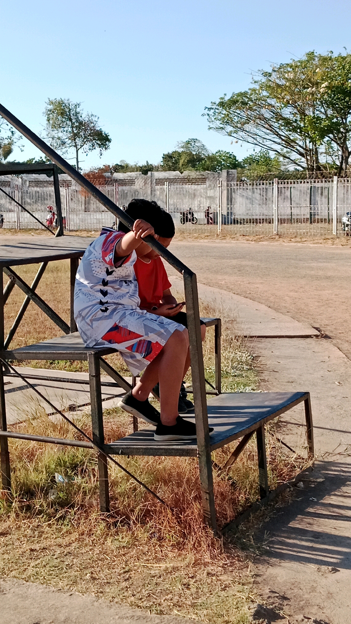Two children are sitting on bleachers outdoors. The child in the foreground is wearing a white shirt with a red, blue, and black geometric pattern and red and blue shorts. They are holding onto a metal railing. The child behind them is wearing a red shirt and black shorts and is looking at a phone. The bleachers are made of metal and wood and are surrounded by dry grass and dirt. In the background, there is a concrete wall with a metal fence in front of it. Trees and power lines are visible beyond the fence. The sky is clear and blue. The image appears to have been taken on a sunny day.