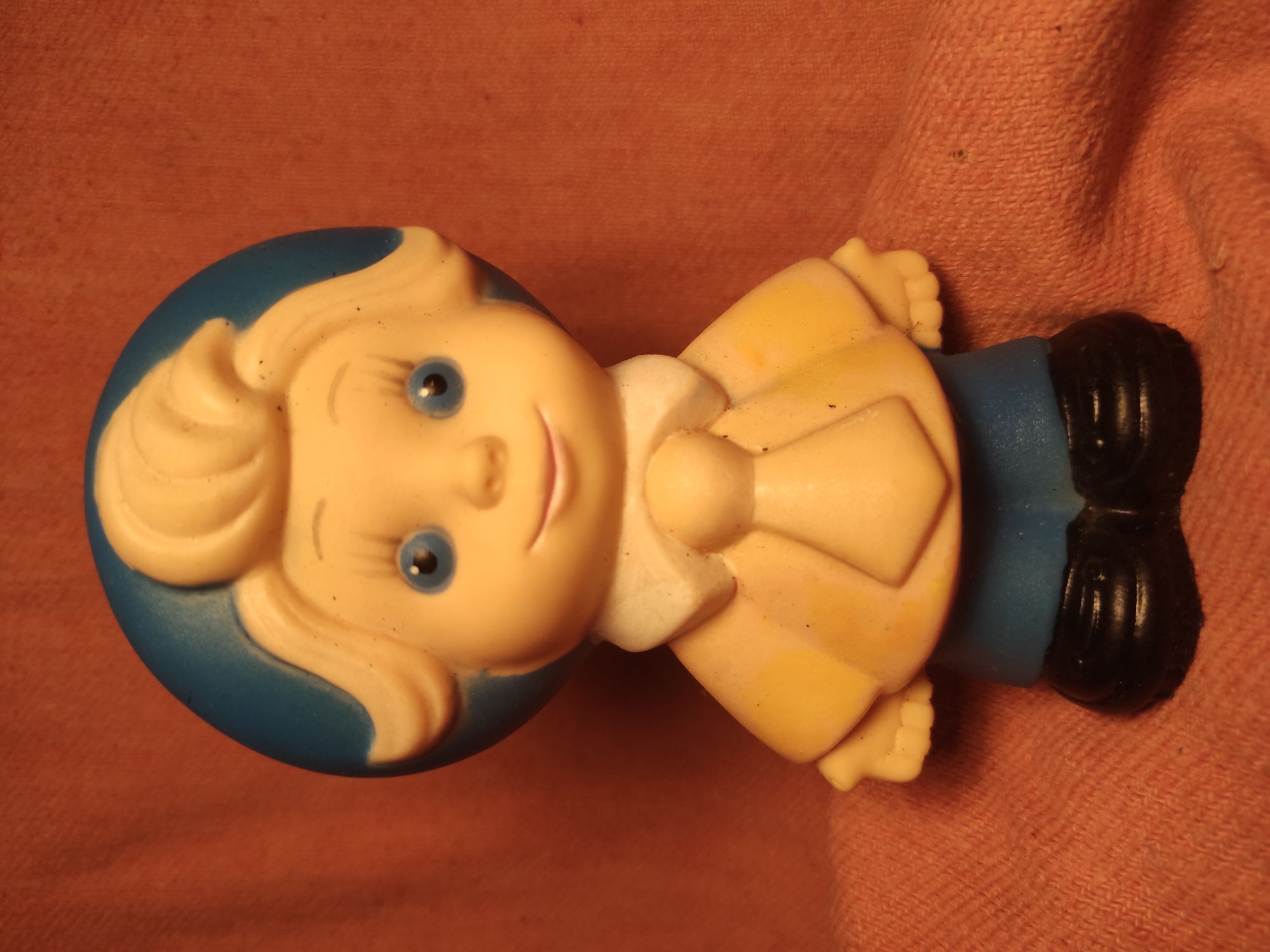 A close-up, head-on studio photograph captures a vintage rubber toy of a cartoonish young boy. The toy stands upright against a textured, salmon-colored fabric background. The boy has a round, pale yellow face with bright blue eyes, dark eyebrows, and a small, smiling mouth. His blonde hair is styled in a swept-back manner, with a distinctive cowlick on top. He wears a blue cap that covers the back of his head. His outfit consists of a yellow shirt with wide lapels, a white collar, and a light yellow tie. Below the shirt, he wears blue pants and black shoes. The toy appears to be made of soft plastic or rubber and shows some signs of age and wear, with visible dust particles on its surface. The lighting is warm and slightly dim, creating soft shadows. There is no information available regarding the location in Chernihiv, Ukraine.