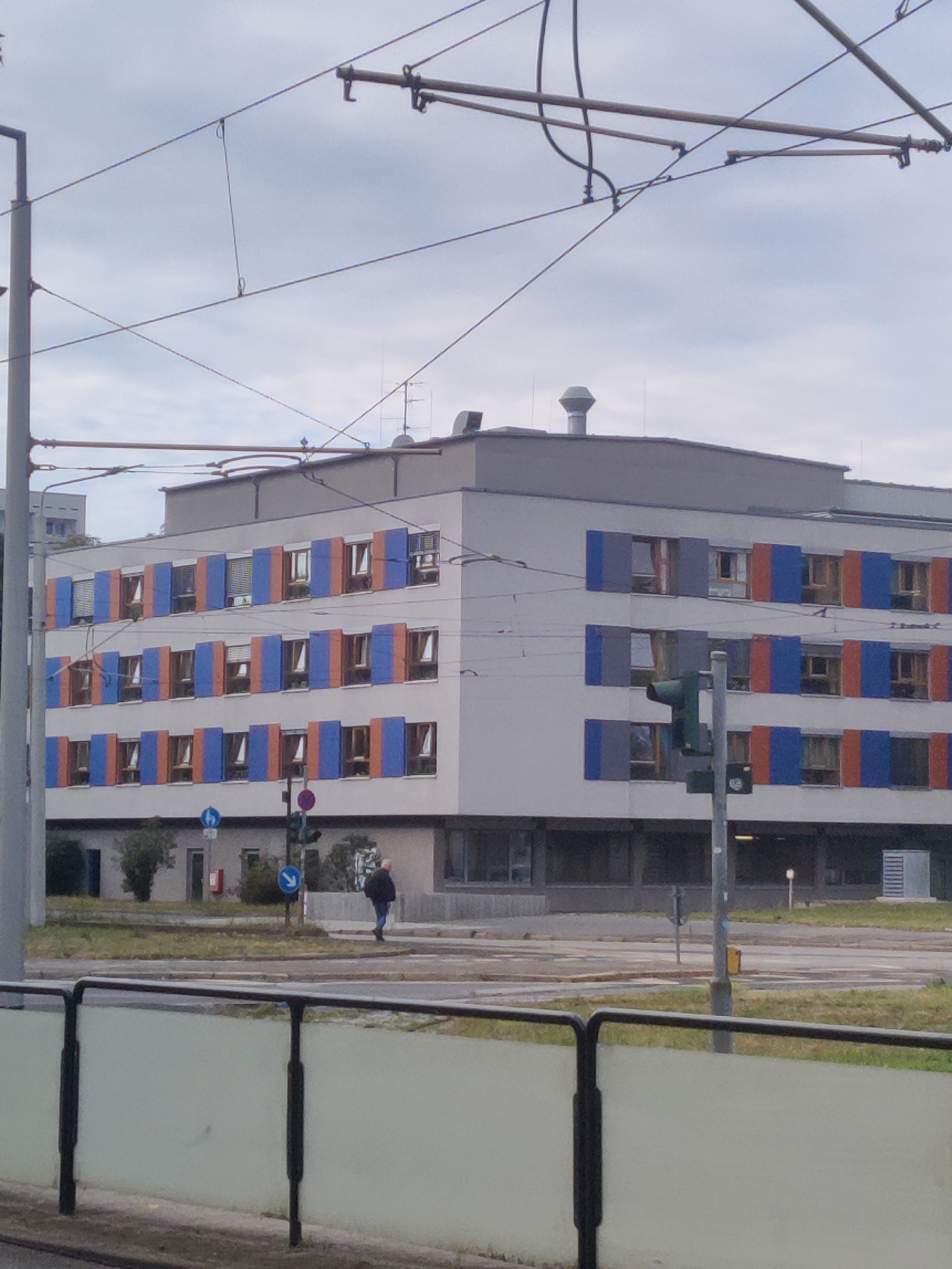 A medium shot captures a multi-story building with a light gray facade and bands of orange and blue shutters covering some of its windows. Overhead, a network of overhead power lines for a tram system crisscrosses the frame against a pale, cloudy sky. In the foreground, a white barrier with black metal supports runs horizontally. A lone individual, seen from behind, walks away from the camera on a street in front of the building. Traffic signals and road signs are visible, suggesting an urban environment. The scene is likely located near the tram lines in Dresden, Germany, based on the presence of the overhead power infrastructure and typical European urban design elements.